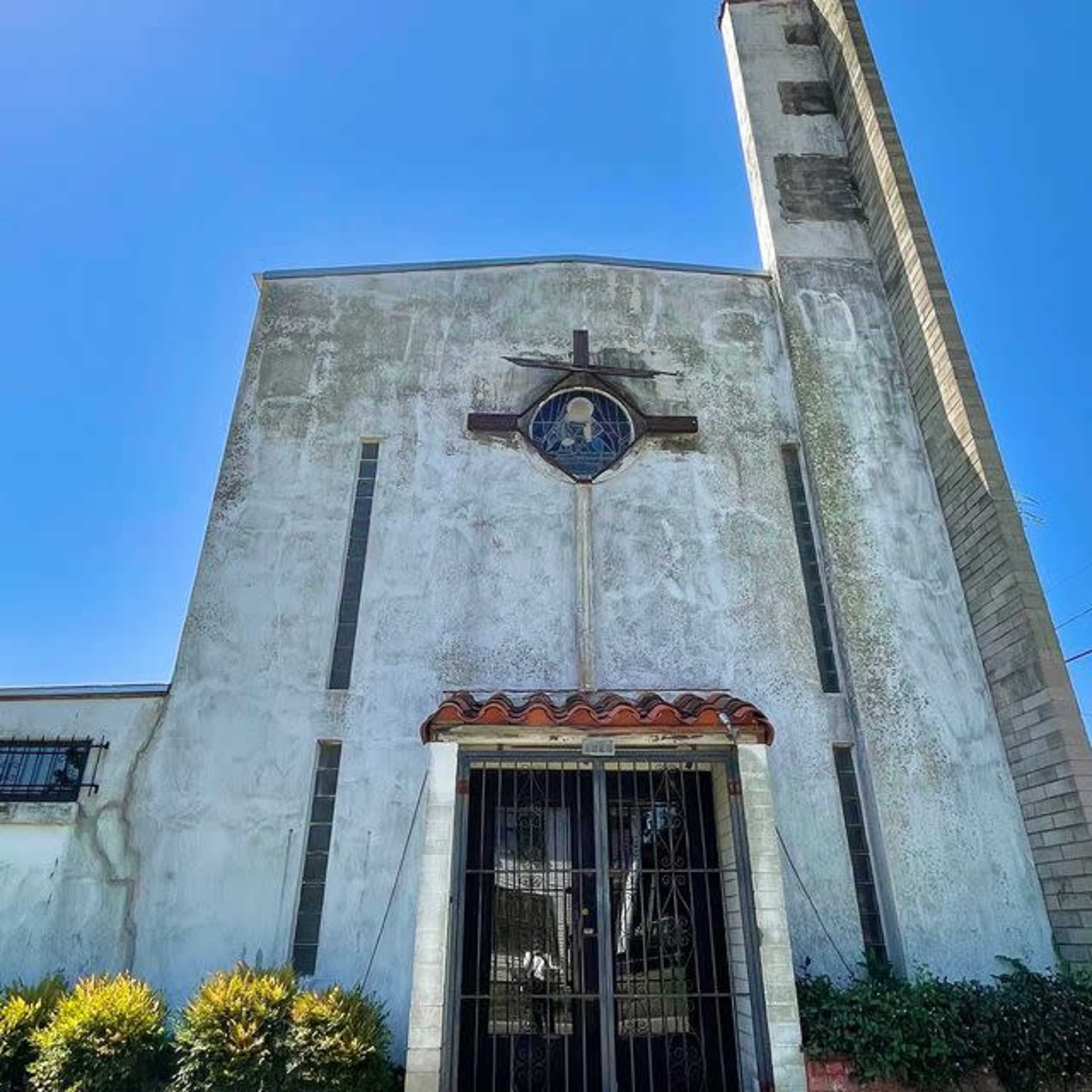A weathered church facade with a gated entrance and a cross above the door, set against a clear blue sky.