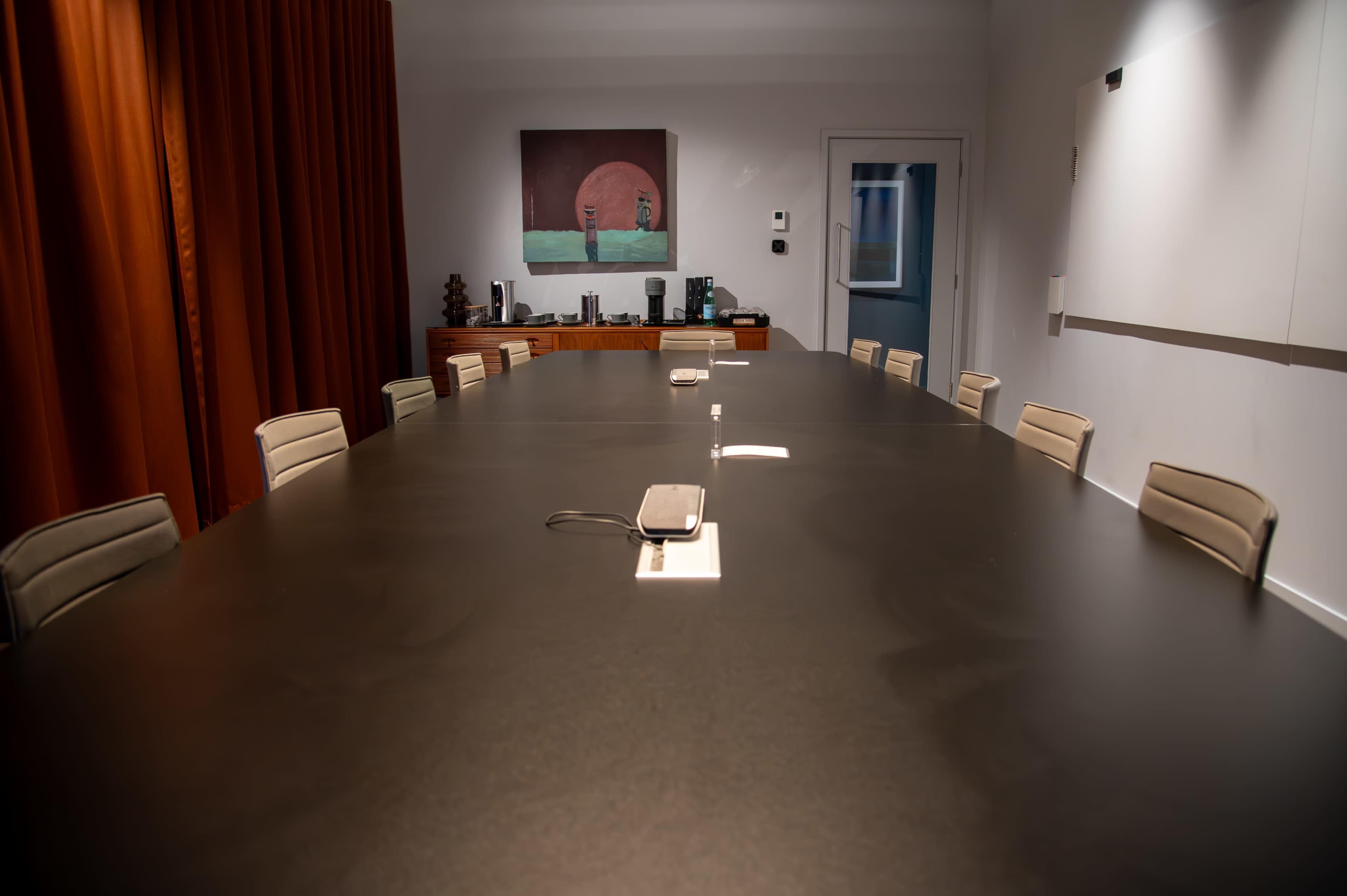 A large conference room features a long black table surrounded by beige chairs, with a painting on the wall and equipment visibly arranged in the background.