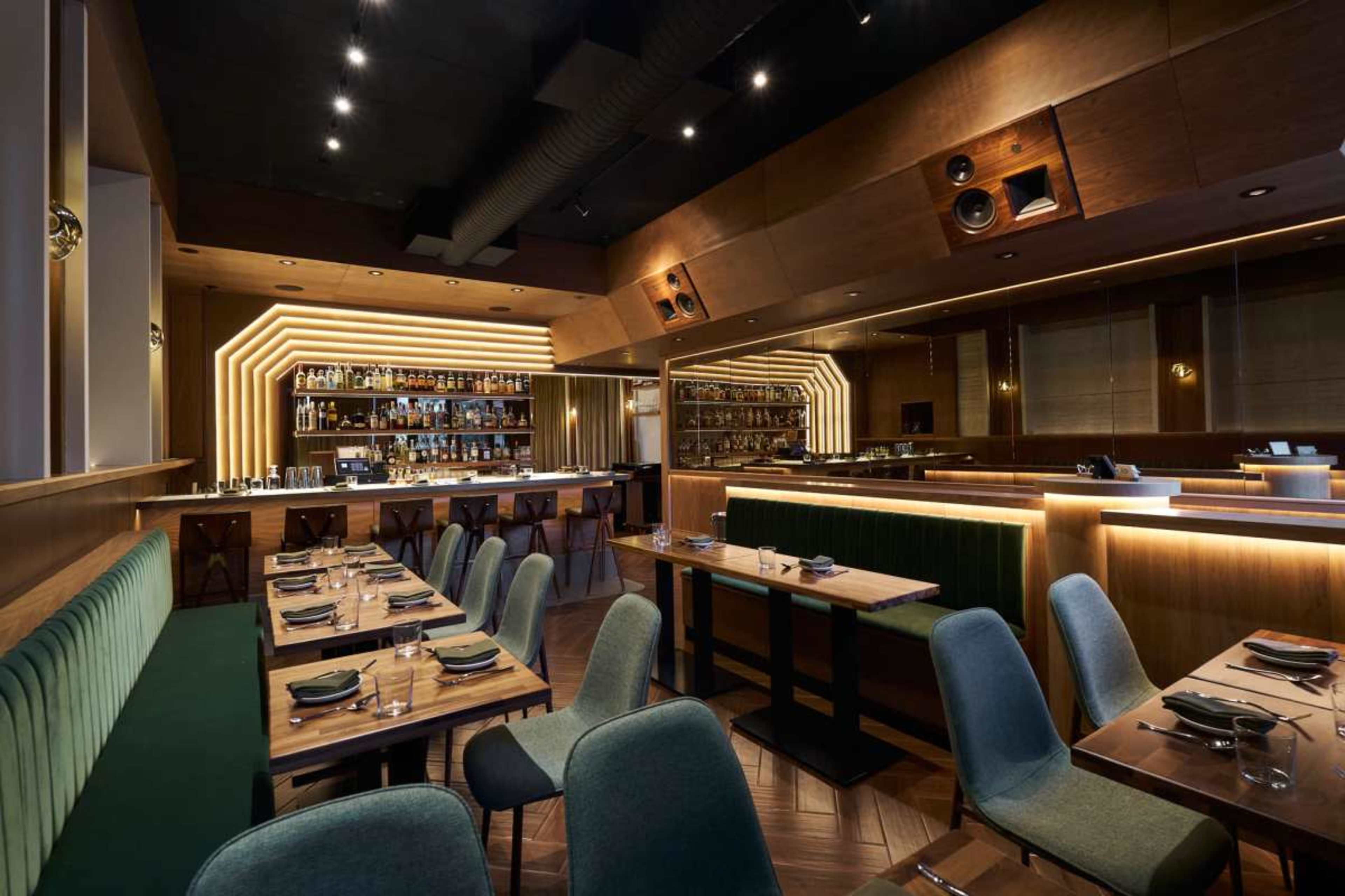 The image shows a modern restaurant interior featuring wooden furnishings, green upholstered seating, and ambient lighting highlighting a bar area.