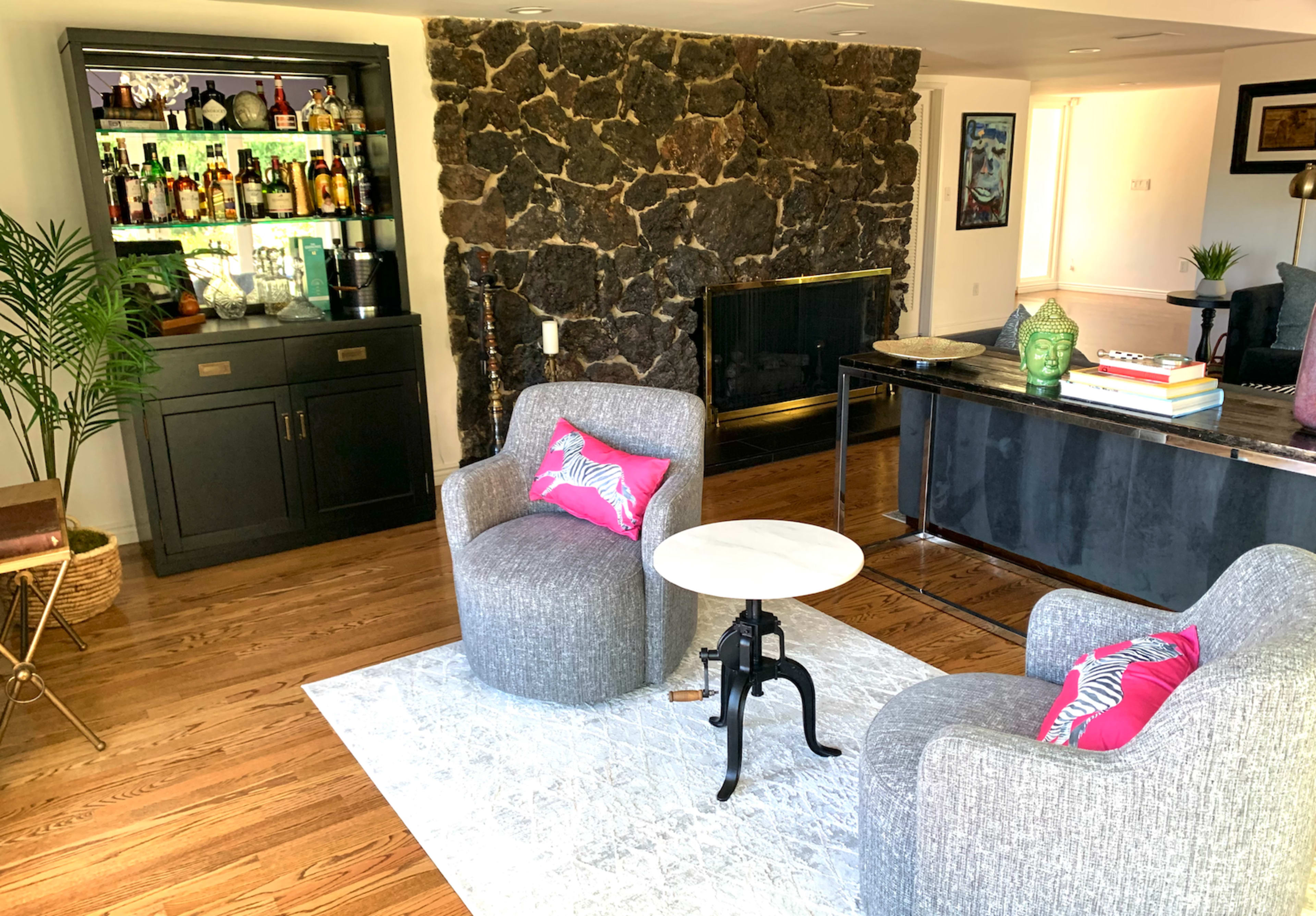 The image shows a living room with two grey chairs and a small round table in front of a stone fireplace, along with a bar cabinet displaying bottles and a decorative plant nearby.