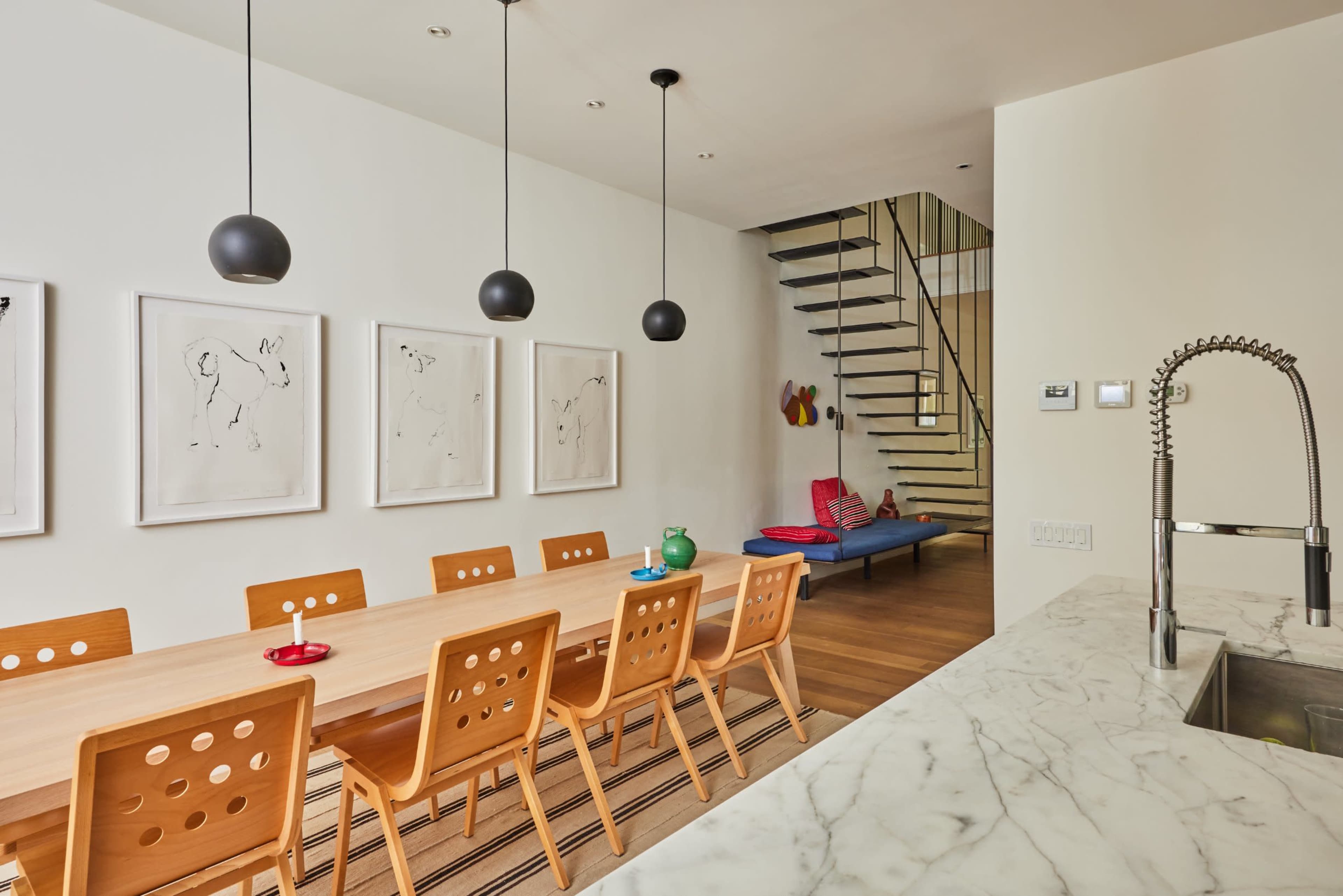 The image depicts a dining area with a long wooden table surrounded by eight chairs, framed artworks on the walls, and a staircase in the background.