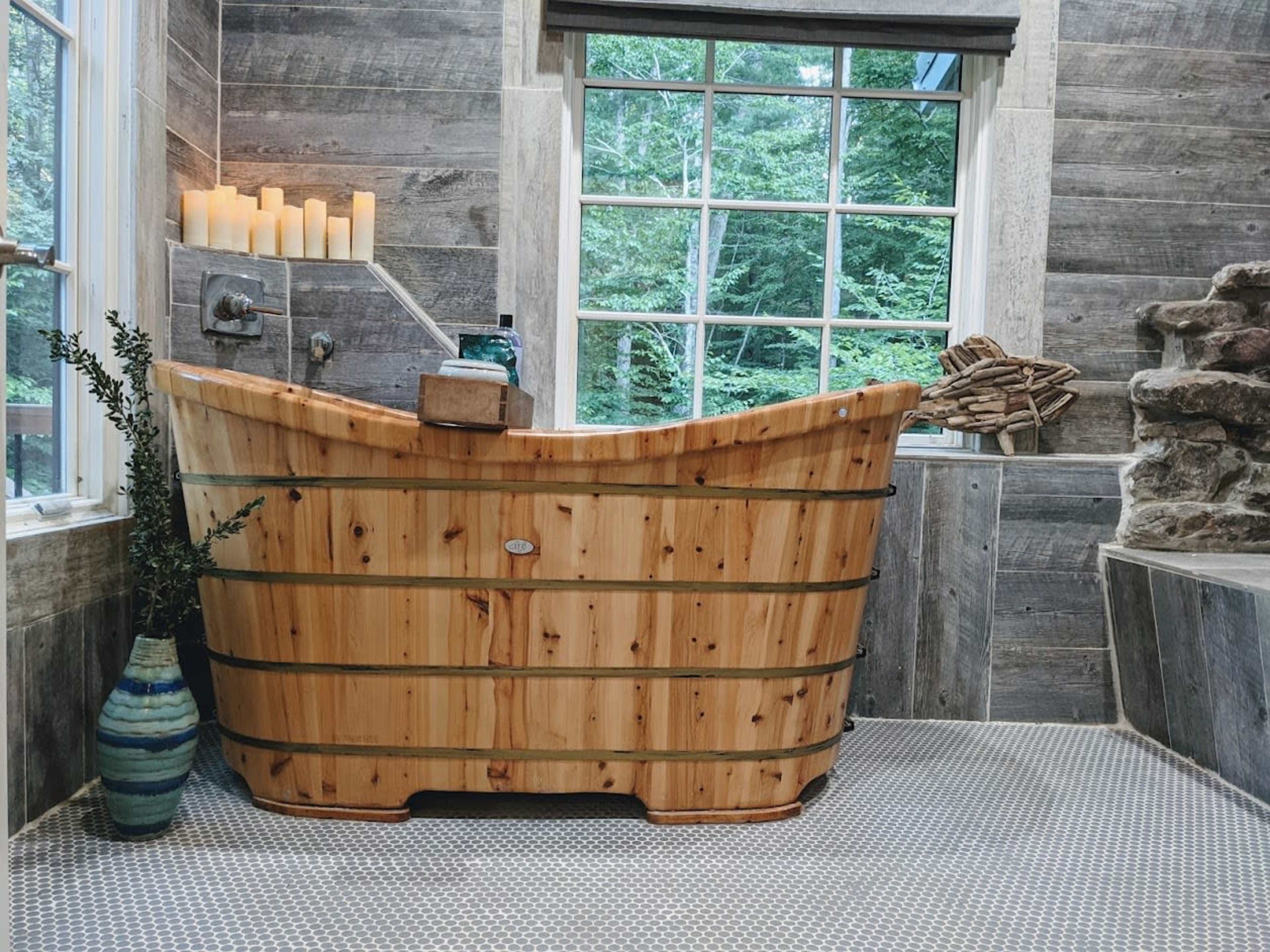A wooden bathtub is positioned in a room with stone walls and a large window overlooking greenery.