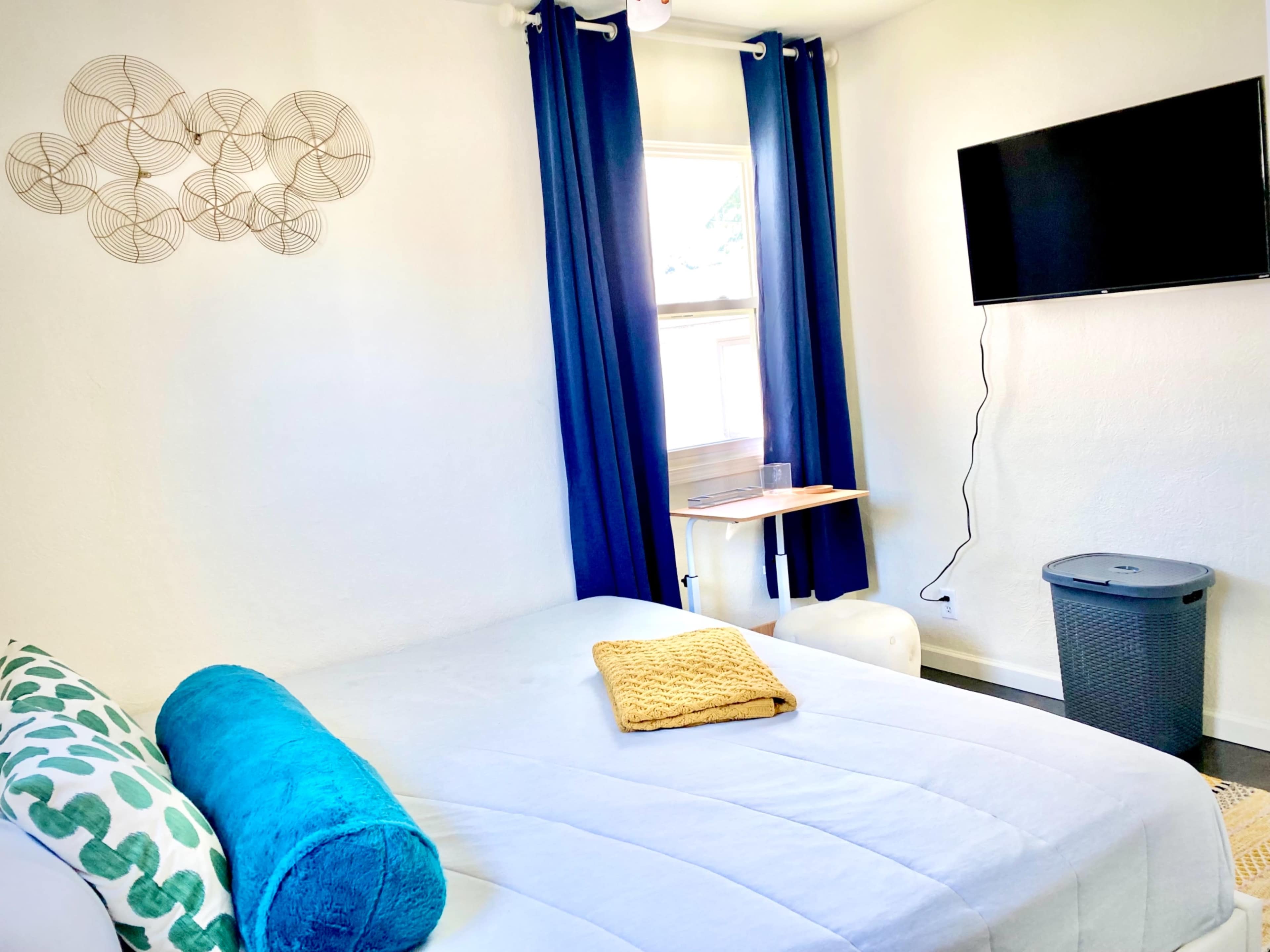 A cozy bedroom with a bed covered in light bedding, a small side table, a TV mounted on the wall, and blue curtains draped over a window.