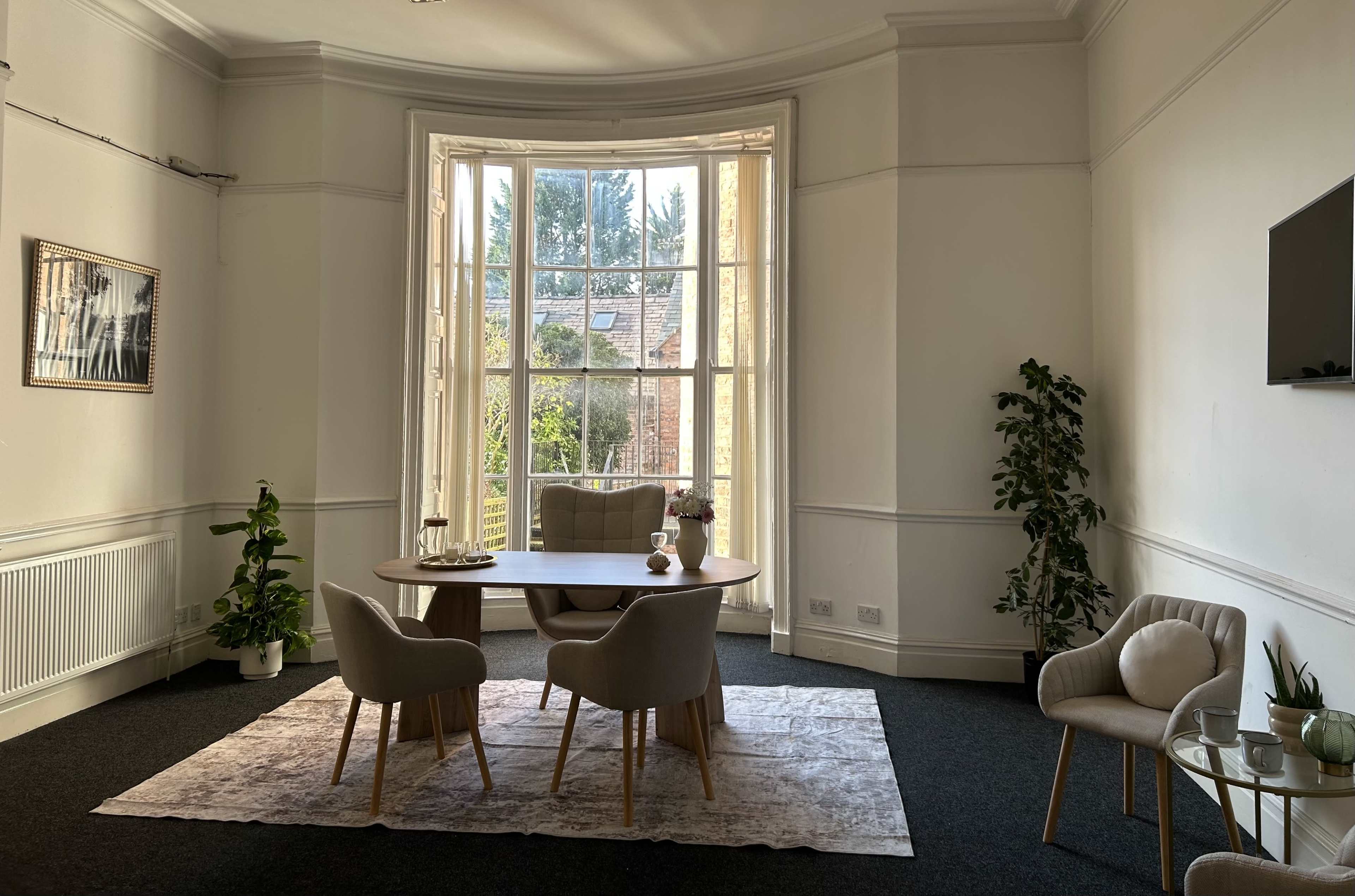 The image shows a bright, airy room featuring a round dining table with four chairs, a large window overlooking greenery, and a few decorative plants.