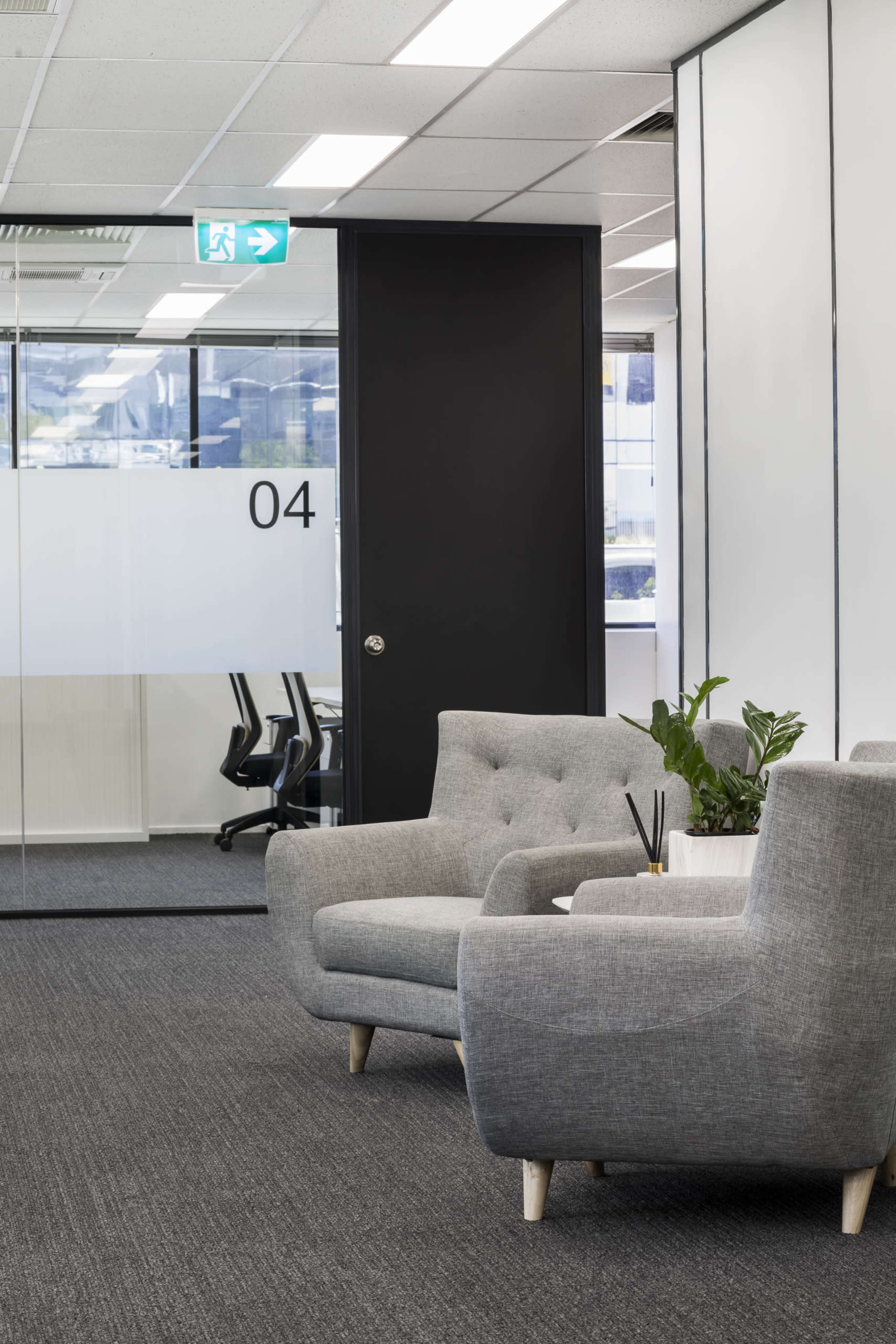 The image shows a modern office interior with two grey armchairs, a small table, and a view of an adjacent meeting room through a glass door marked with the number 04.
