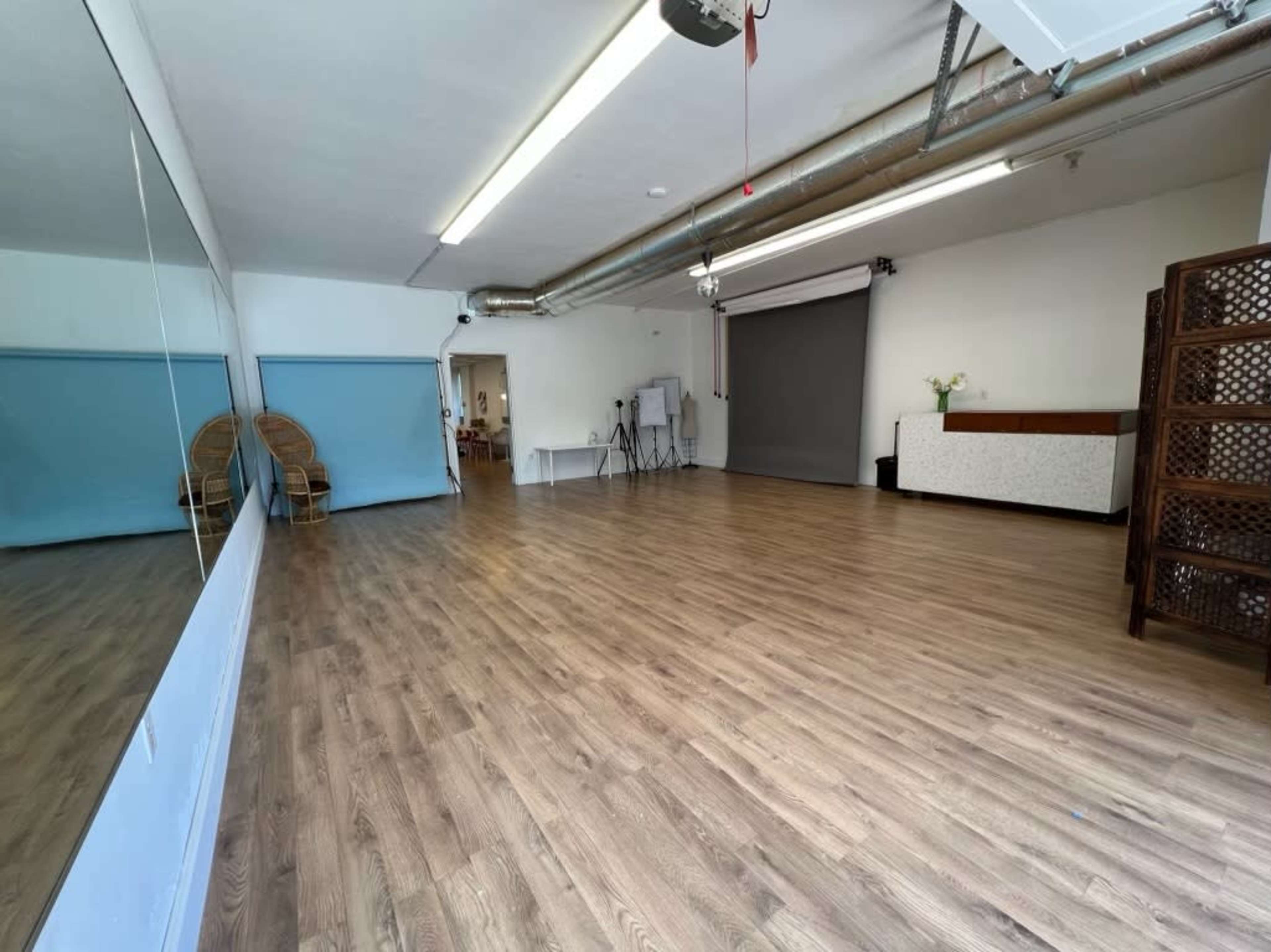 A spacious, empty room with light wood flooring, a large mirror along one wall, and a folding partition in the back.