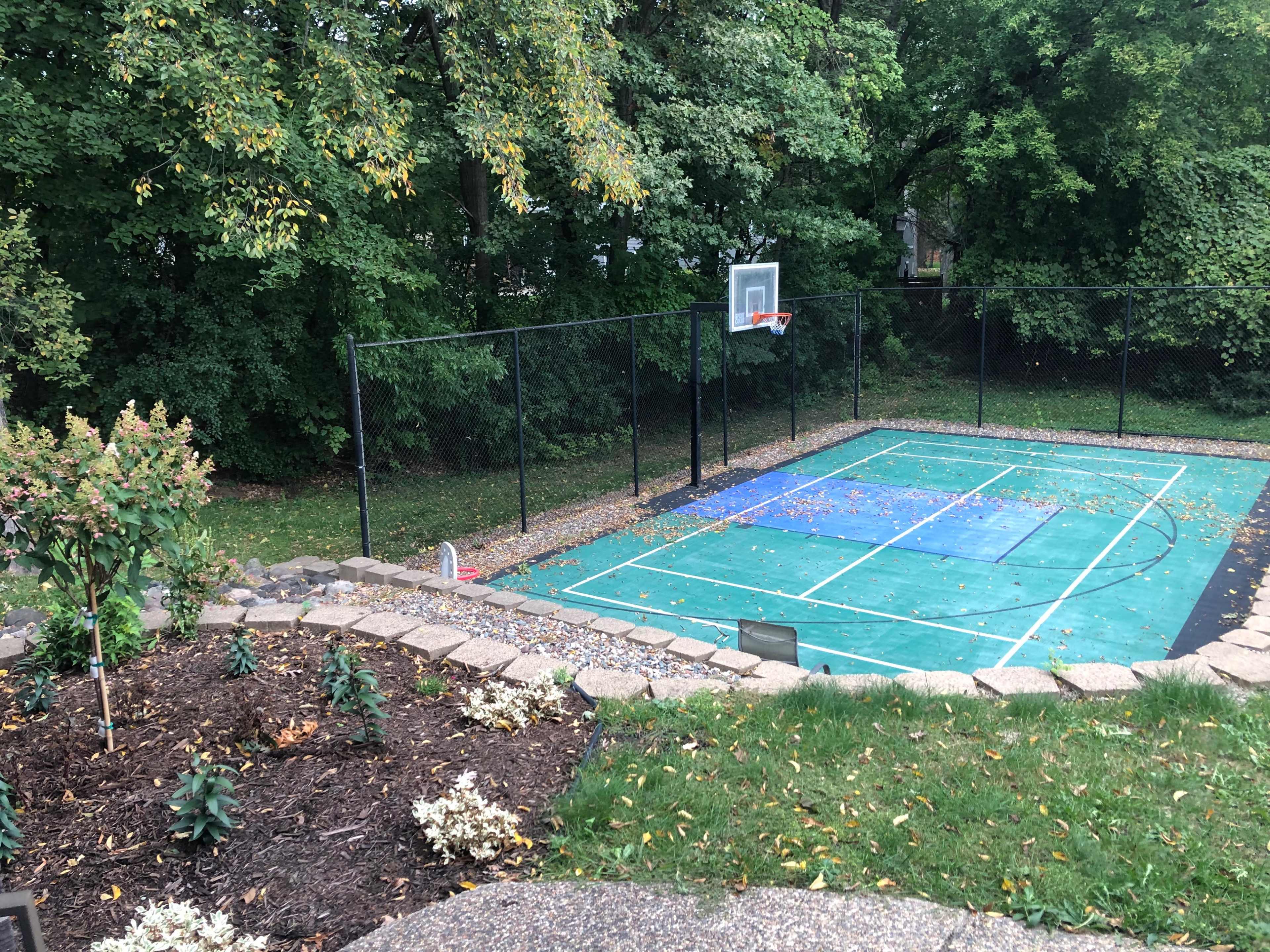 A basketball court surrounded by a chain-link fence, with a garden and lawn area in the foreground.