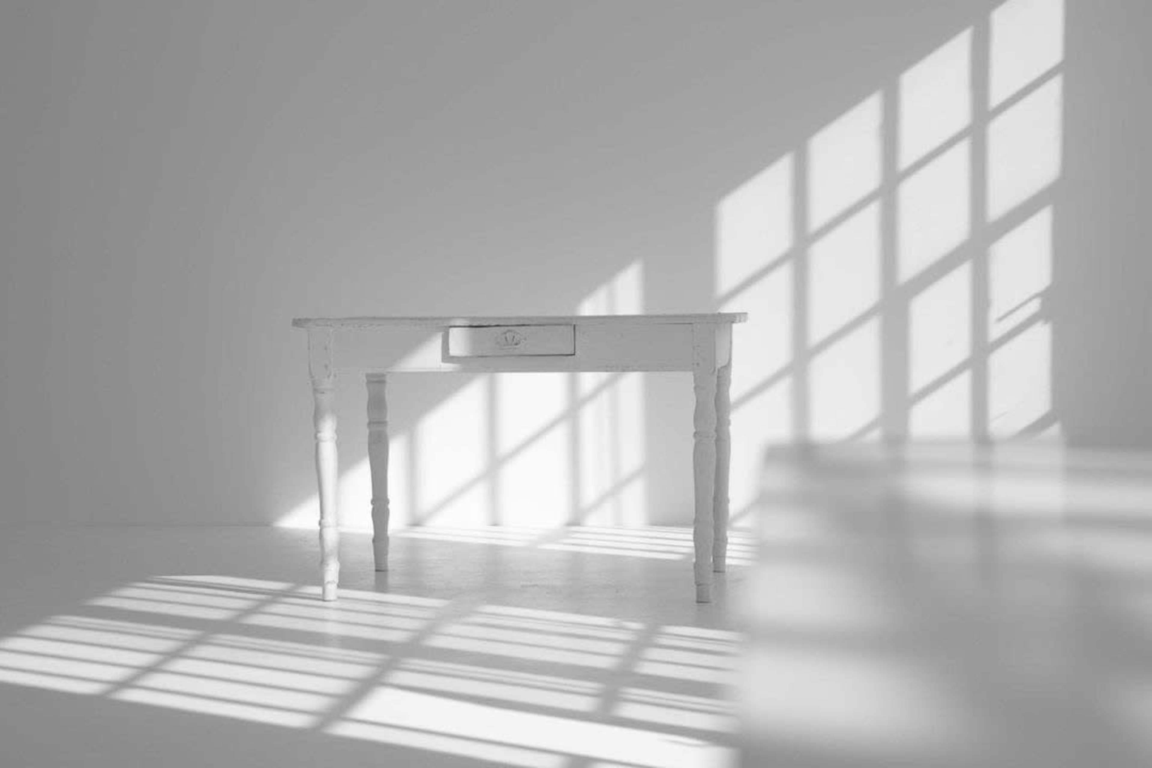 A white table with a single drawer casts shadows on a light-colored floor, illuminated by sunlight streaming through a window.