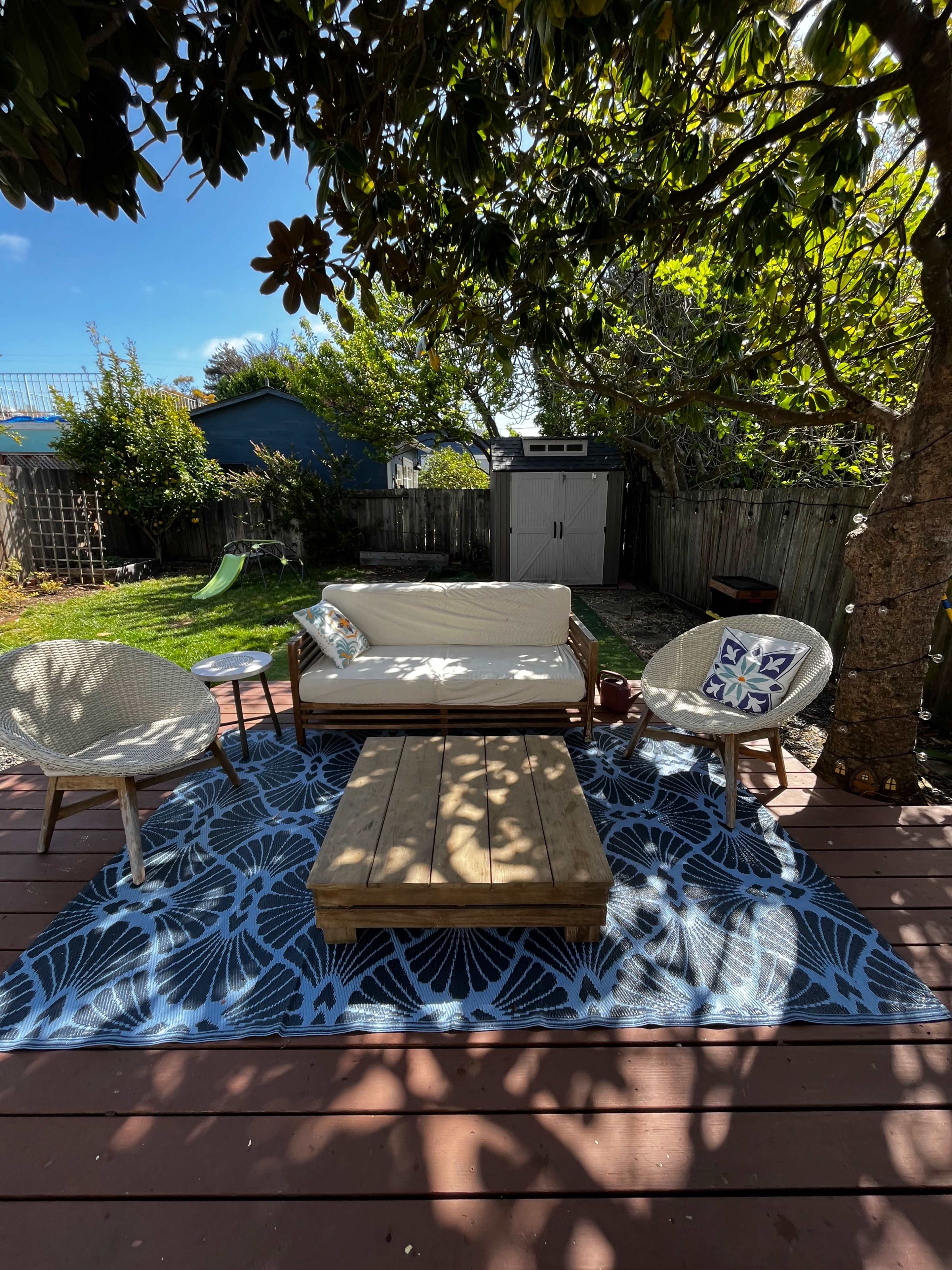 A patio area features a low wooden coffee table, two chairs, and a couch arranged on a patterned rug under a tree.
