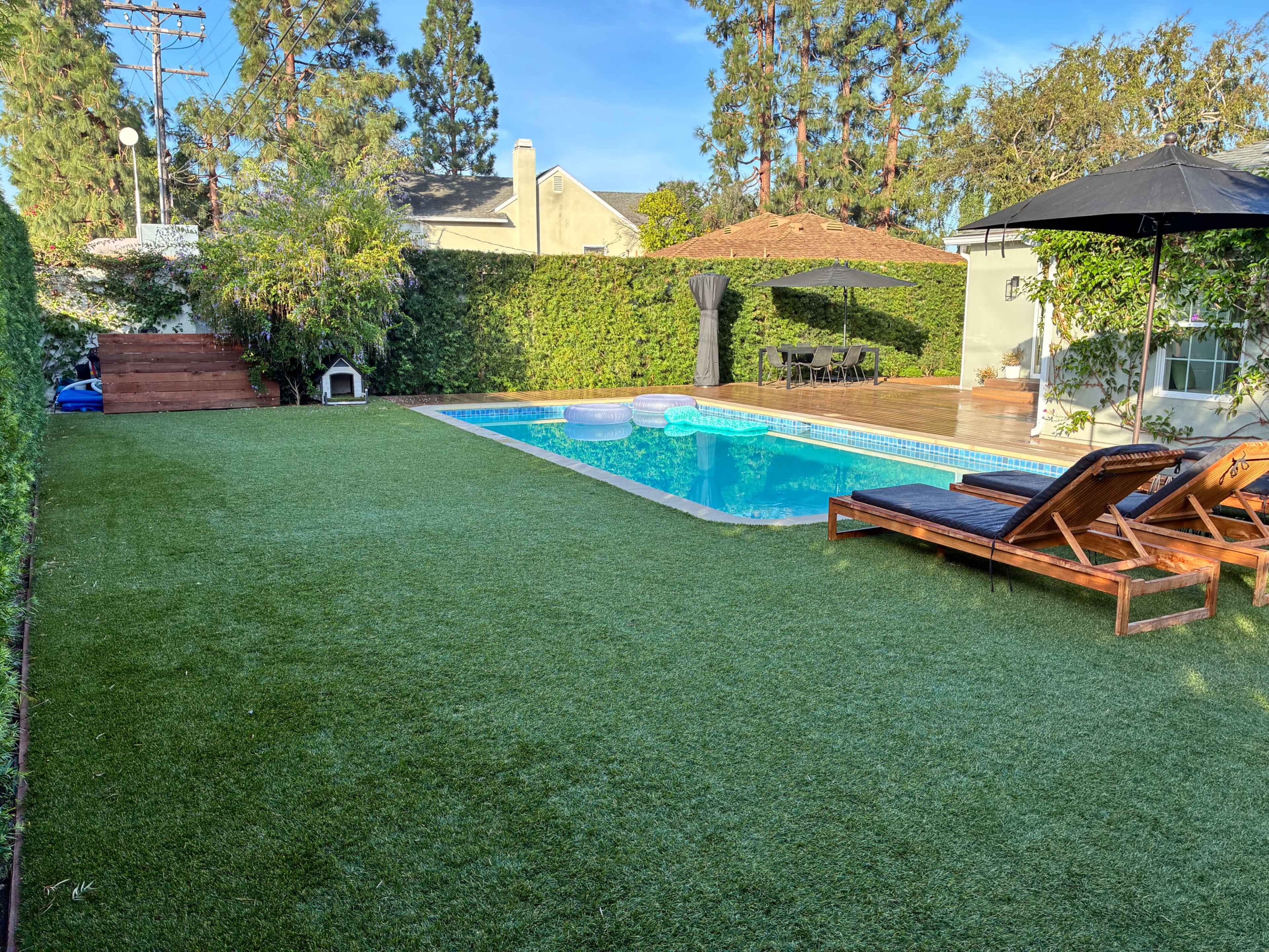 The image shows a backyard featuring a swimming pool surrounded by artificial grass, lounge chairs, and lush greenery.