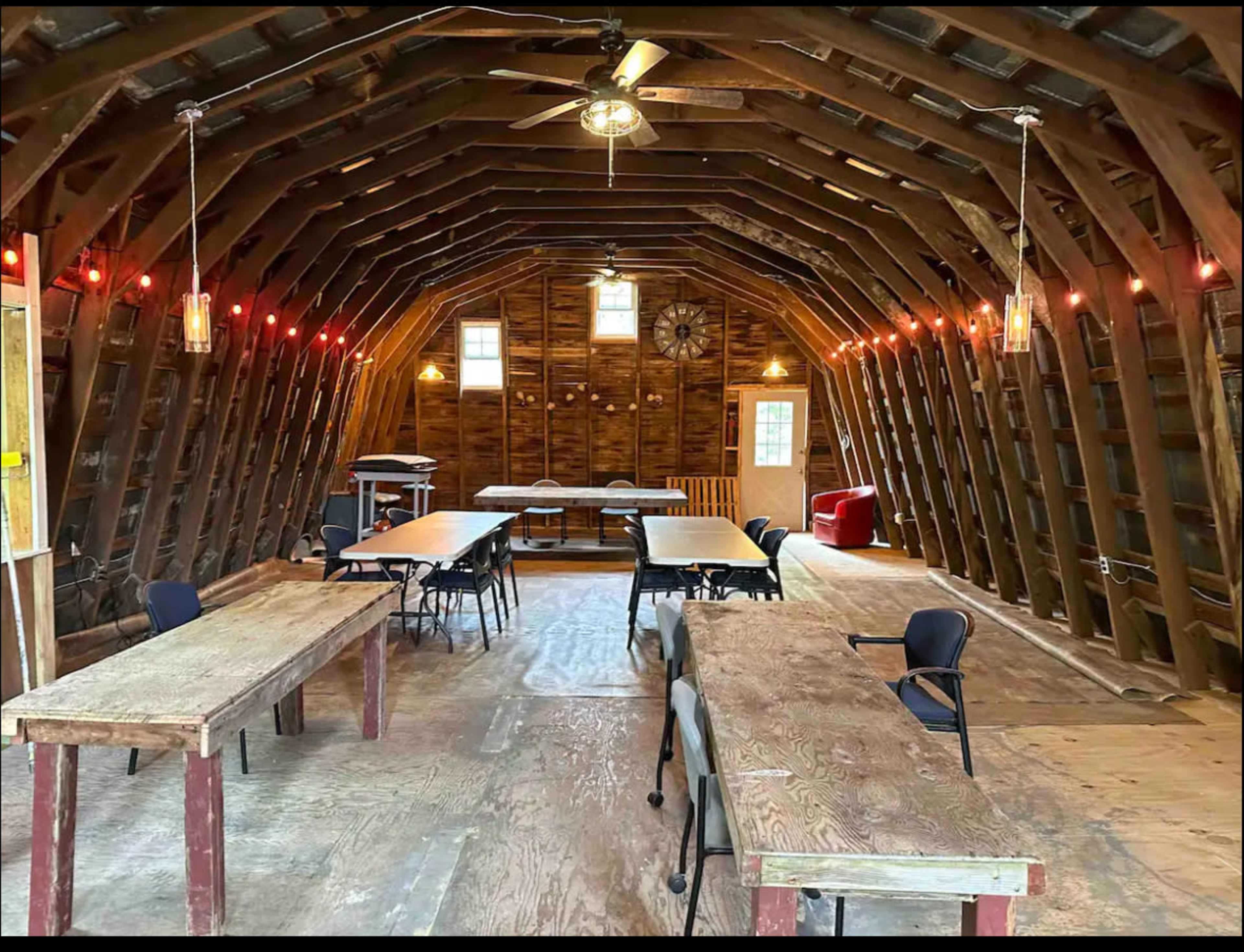 The interior of a rustic barn features several long tables and chairs arranged in rows, with warm lighting and wooden beams visible above.