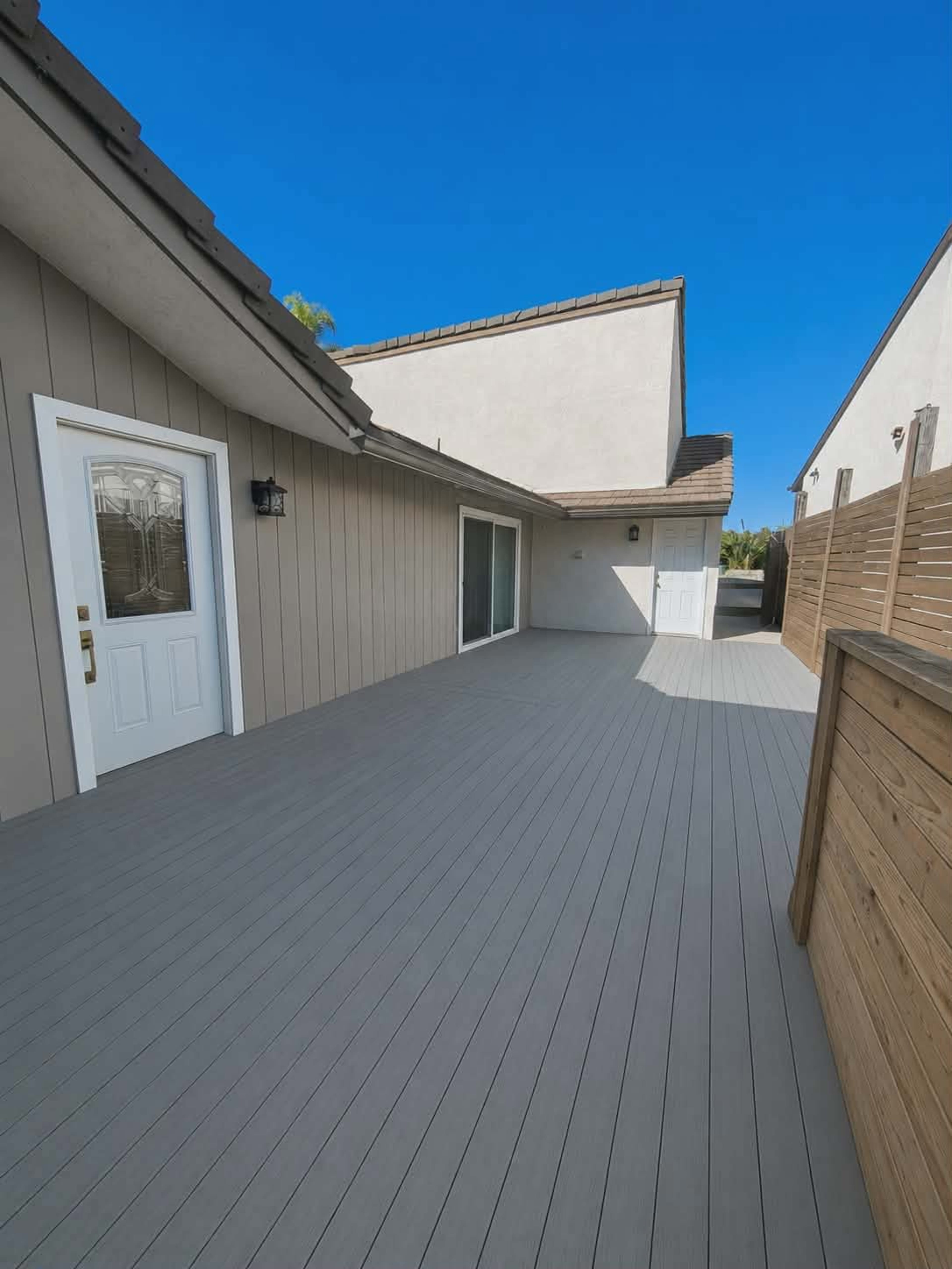 The image shows a spacious wooden deck extending along the side of a house, flanked by walls and with clear blue skies above.