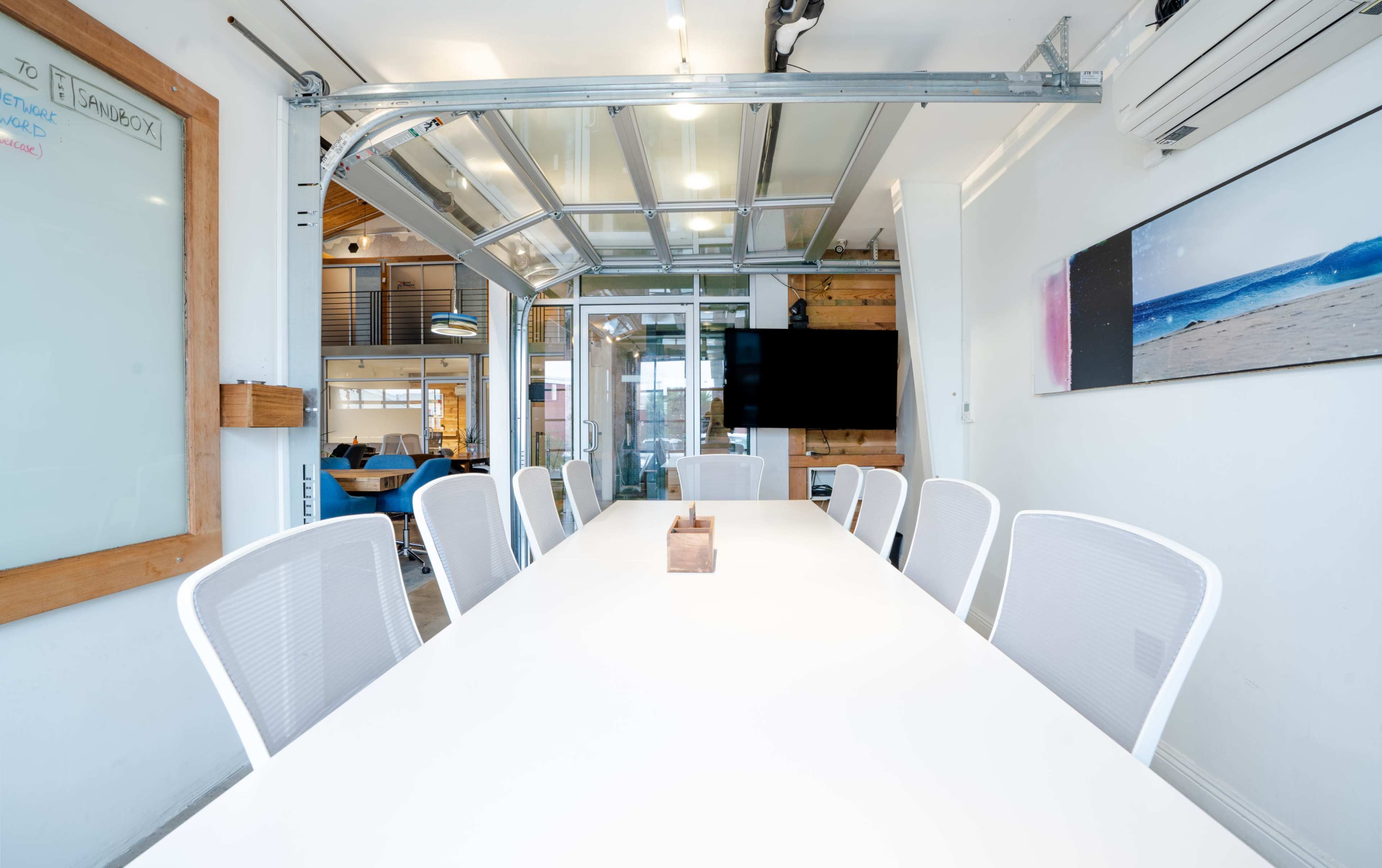 The image shows a modern conference room with a long white table, several white chairs, and large windows letting in natural light.