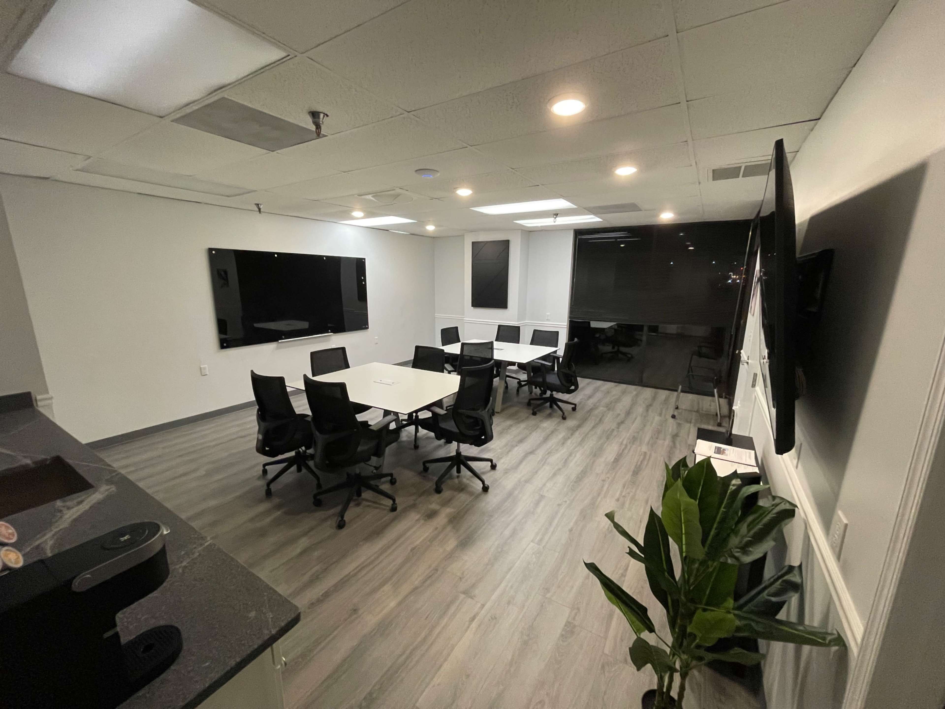 A modern conference room with a large table surrounded by black chairs, multiple screens mounted on the walls, and a green plant in the corner.