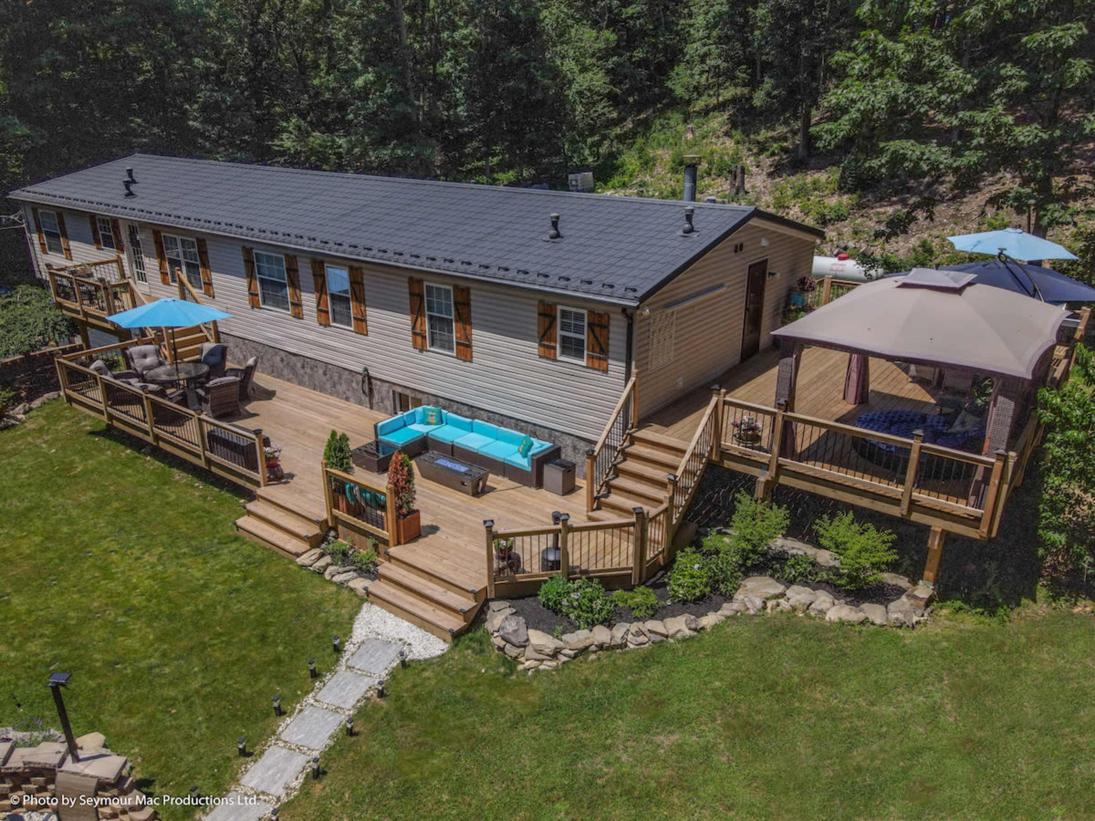 A single-story house with a deck and patio area surrounded by trees in a grassy yard.