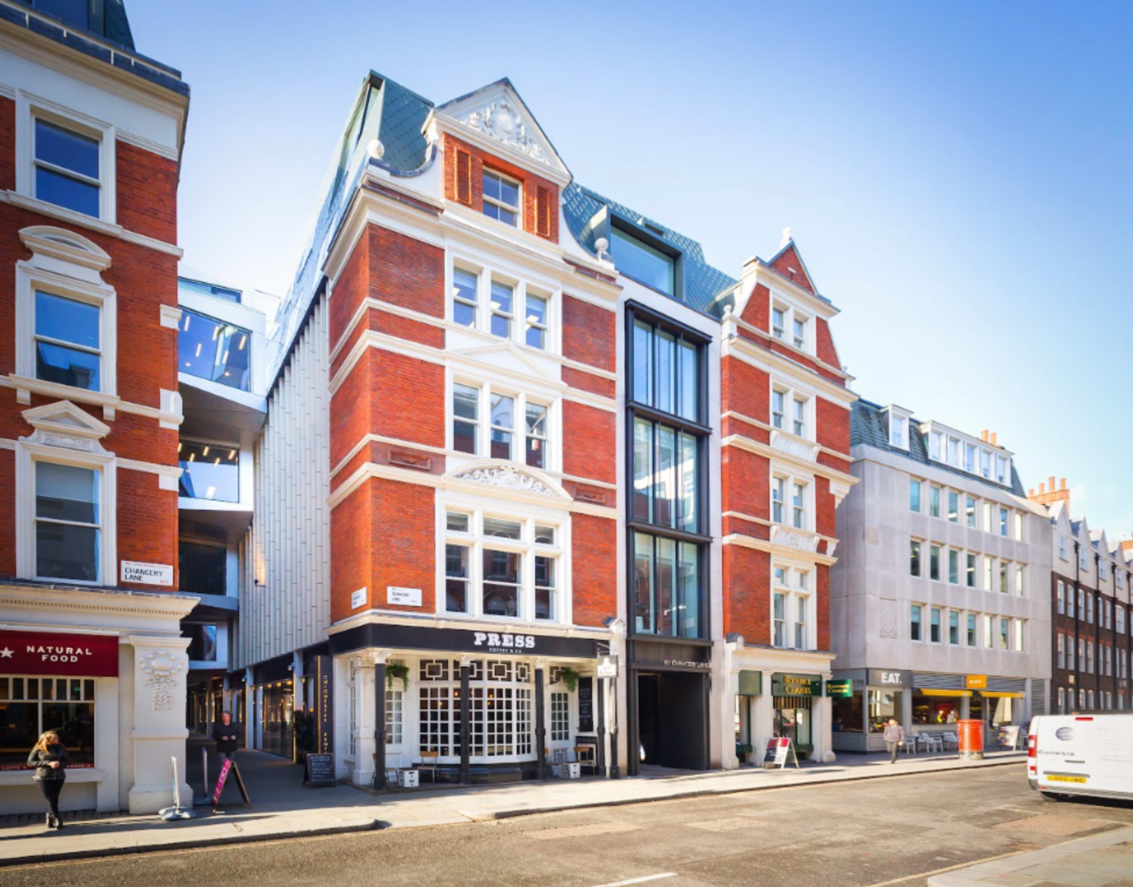 A four-story red brick building with large windows and a modern glass entrance, located on a city street with other buildings visible on either side.