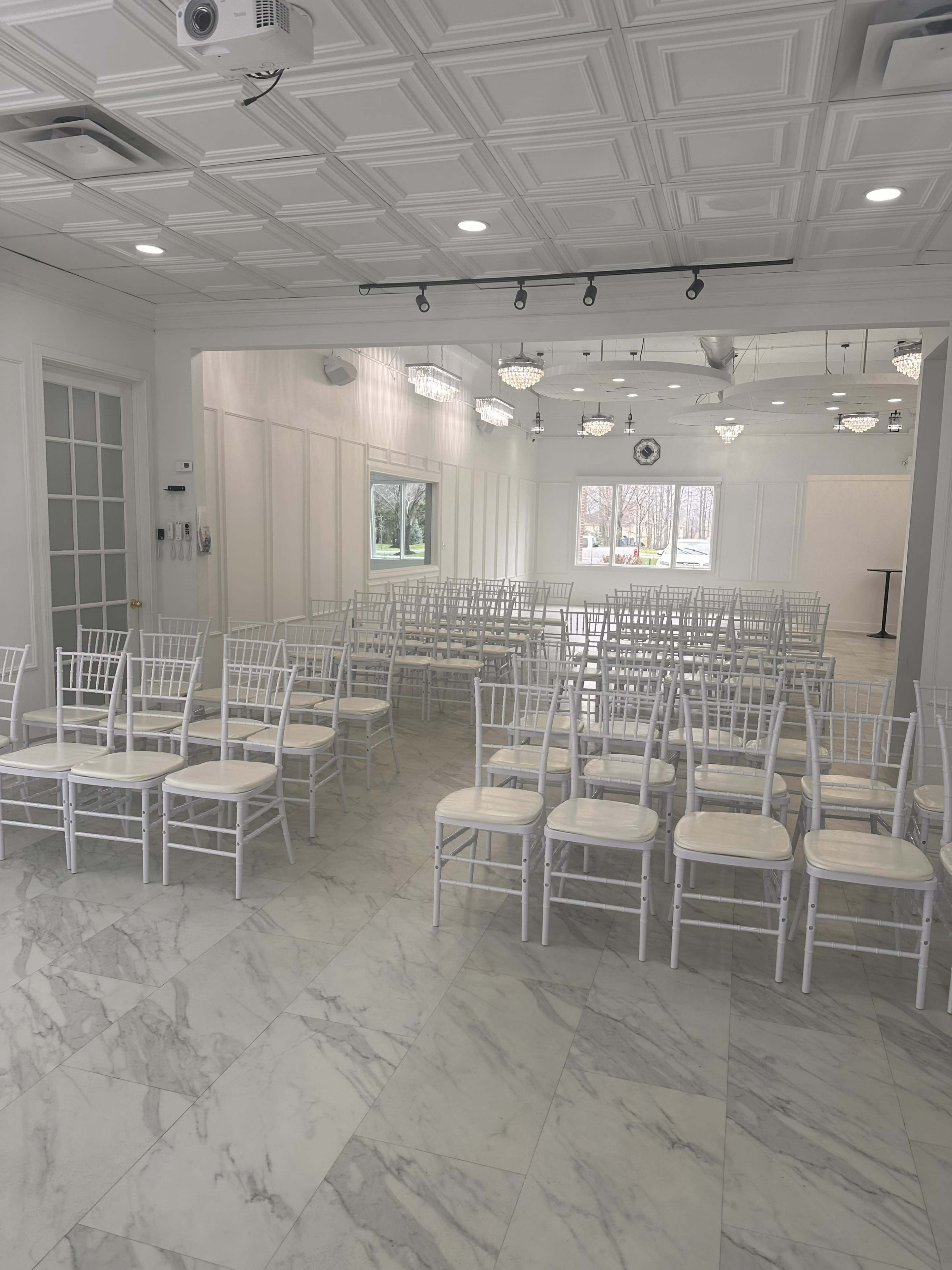 A spacious room features rows of white chiavari chairs arranged on a marble floor, with modern lighting and large windows allowing natural light.