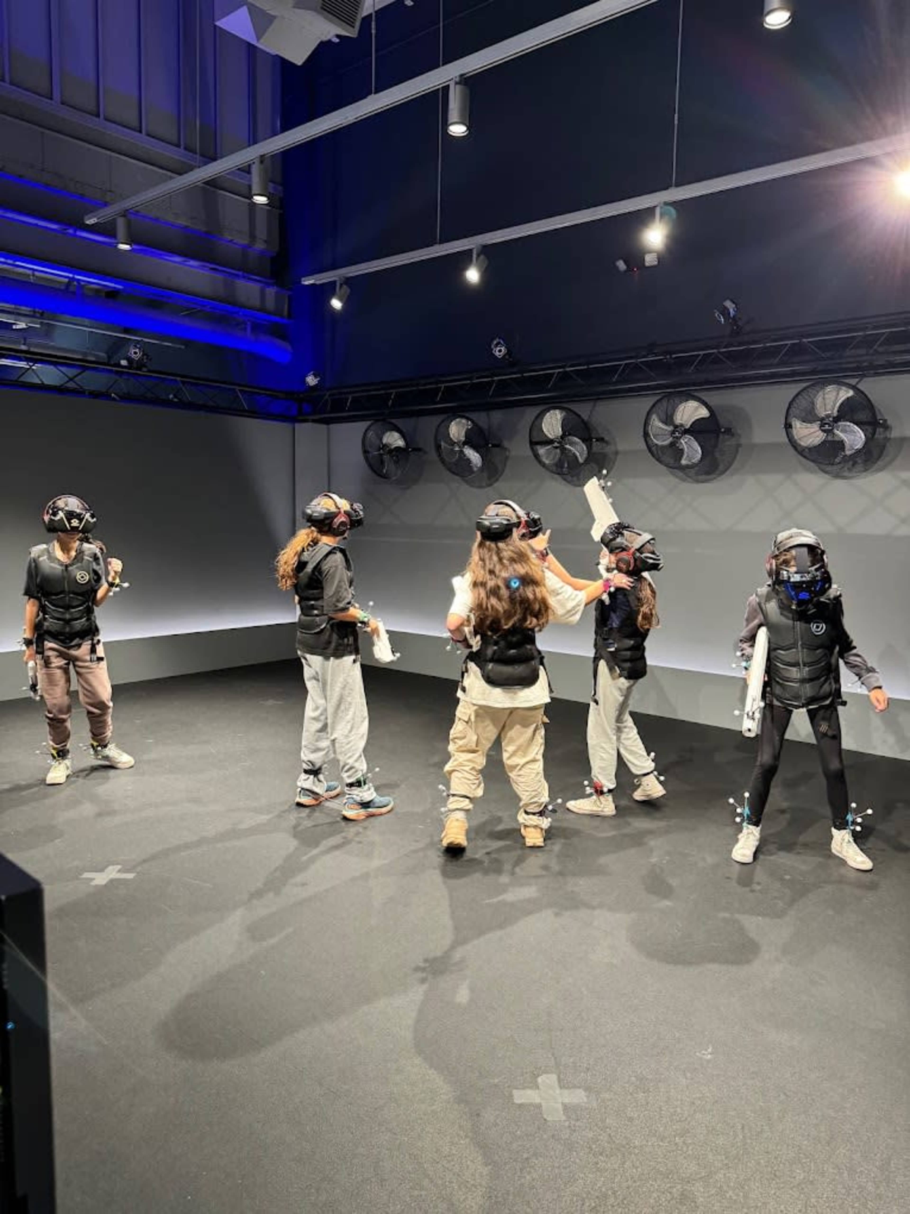 Five individuals wearing virtual reality headsets and gear are standing in a dimly lit room with large fans and black walls.