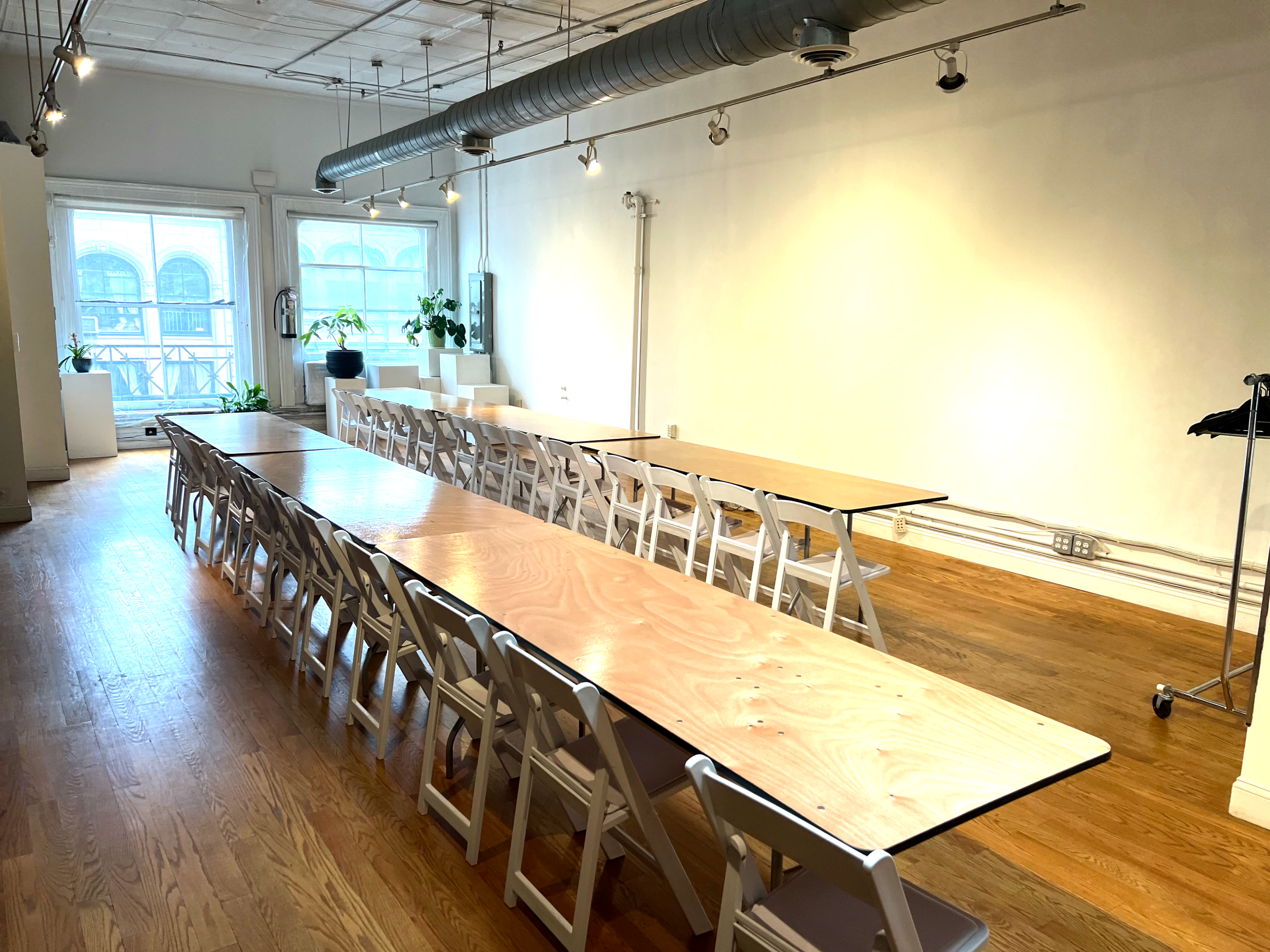 The image shows a spacious room with wooden floors, featuring long tables set up in rows and chairs arranged around them, with large windows allowing natural light to enter.