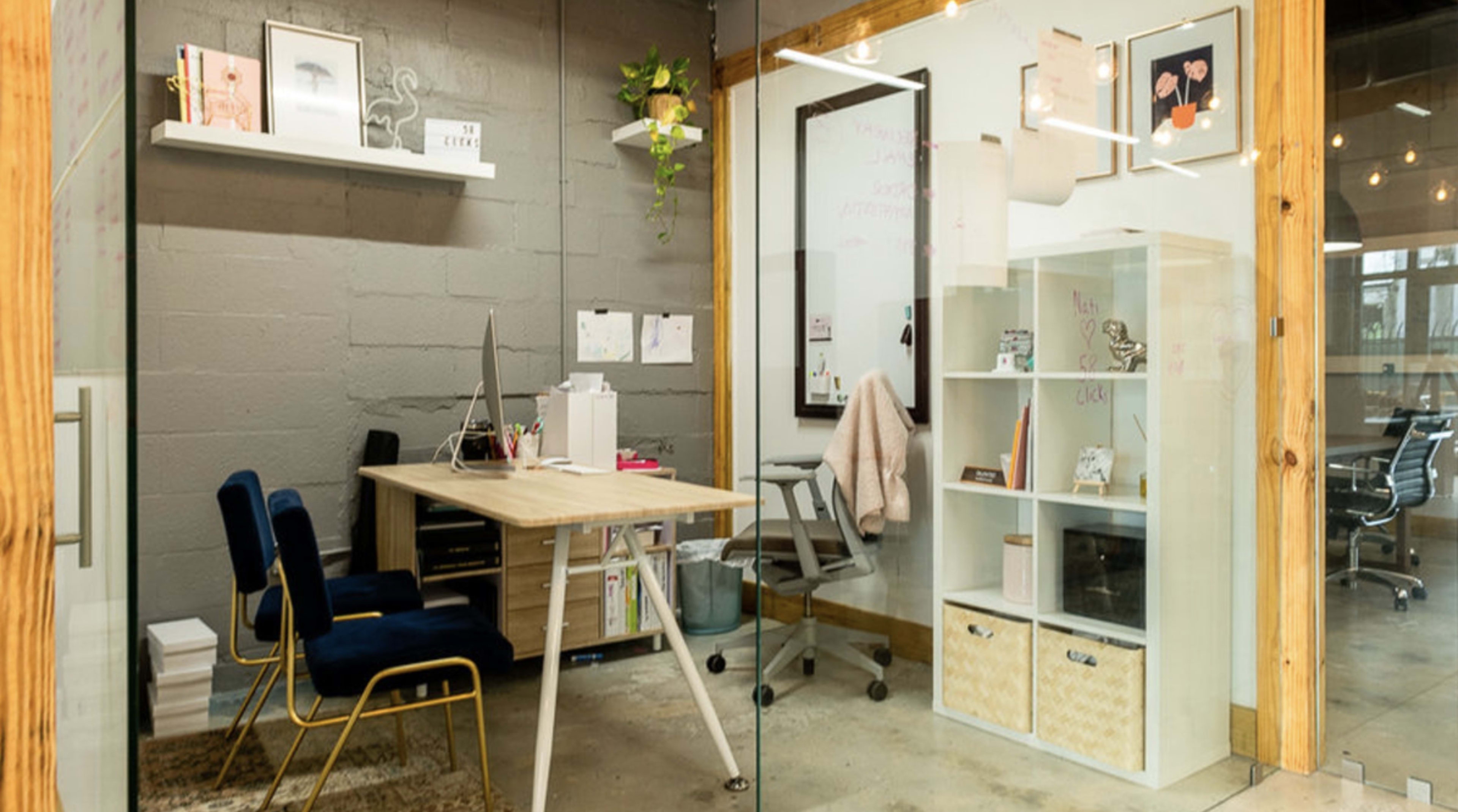 A small office space features a wooden desk with two blue chairs, a computer, and a display cabinet, surrounded by gray walls and glass partitions.