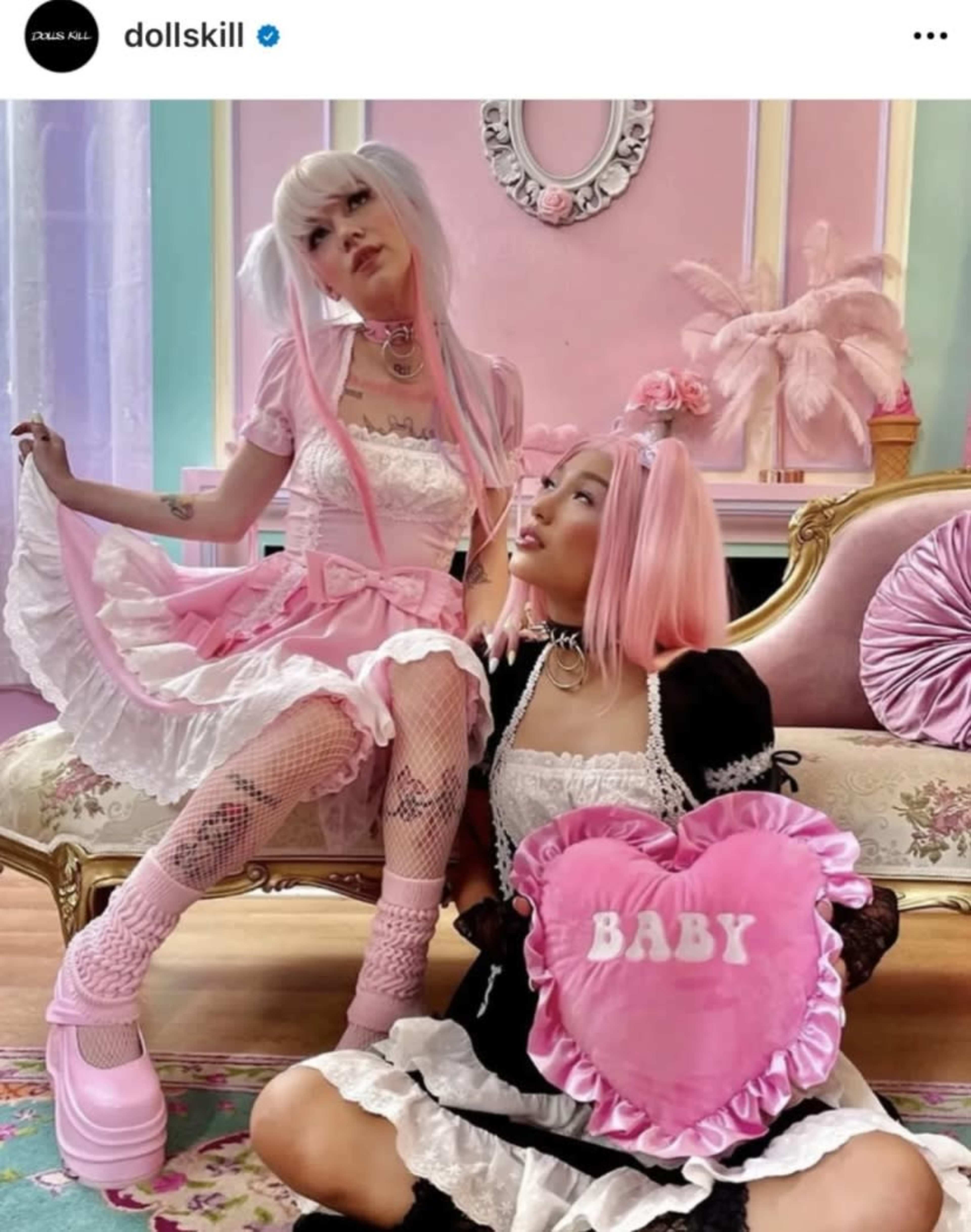 Two women in colorful, frilly outfits are posing on a vintage couch adorned with pastel decor, with one holding a pink heart-shaped pillow that says "BABY."