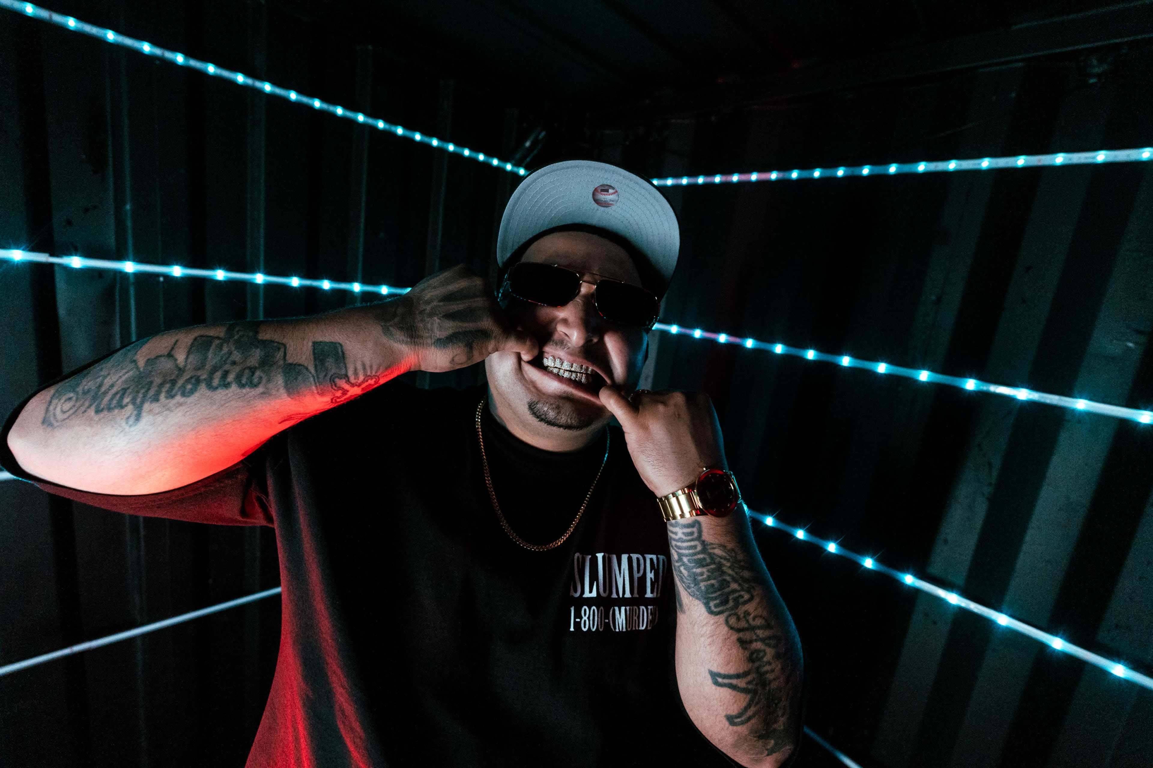 A person with sunglasses is posing inside a neon-lit space, making a gesture that involves his teeth while wearing a black t-shirt and a cap.