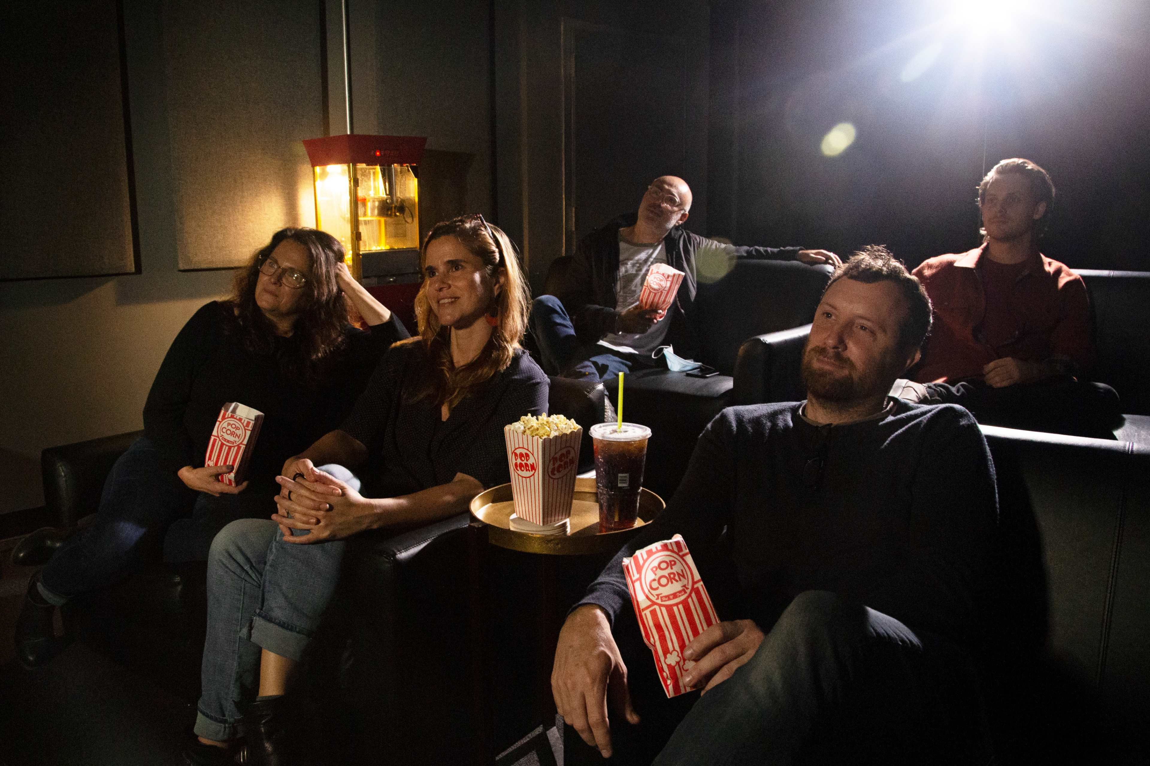 A group of five people sits in a darkened movie theater, enjoying popcorn and drinks while watching a film.