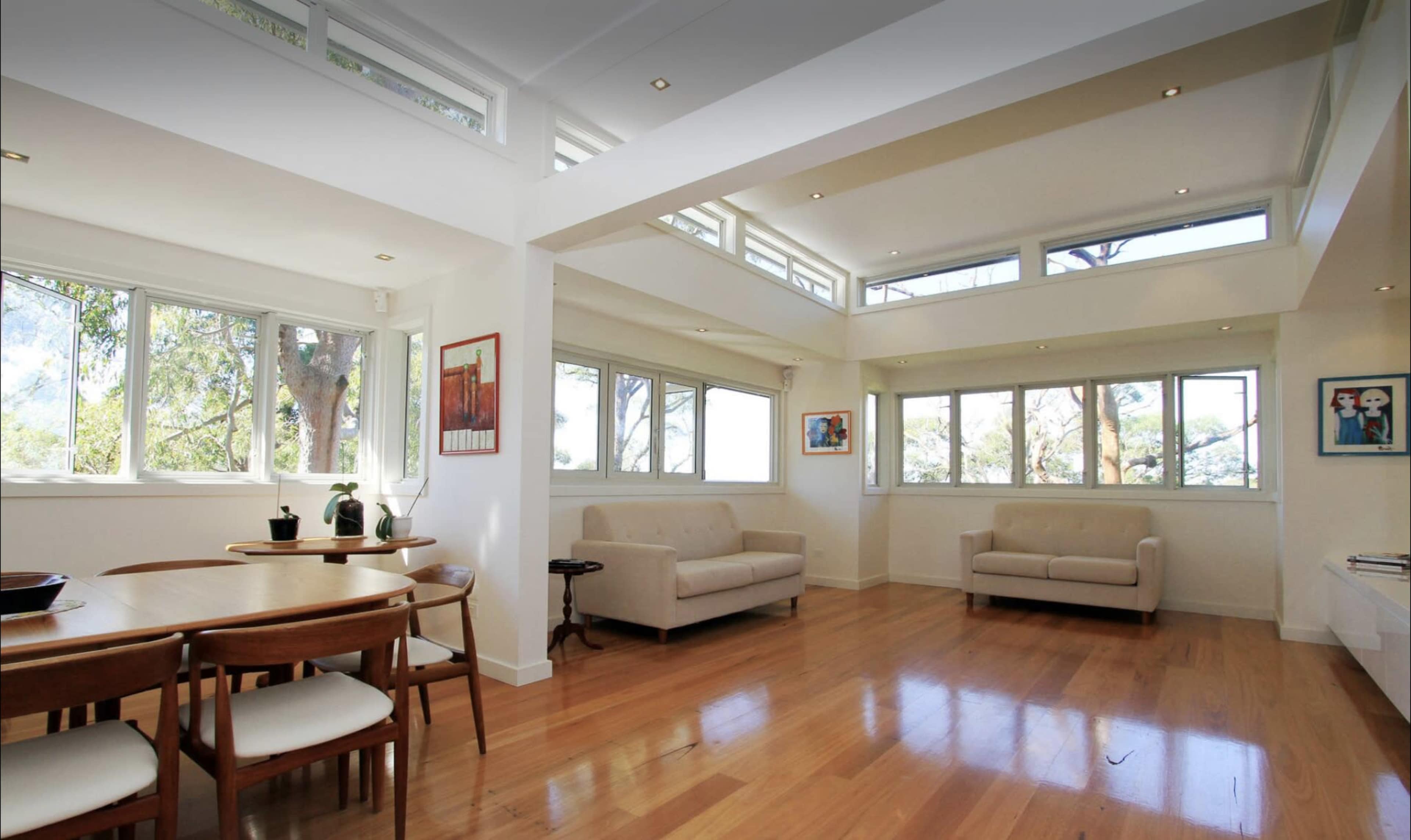 The image shows a bright, open living area with large windows, hardwood floors, and a dining table set for four.