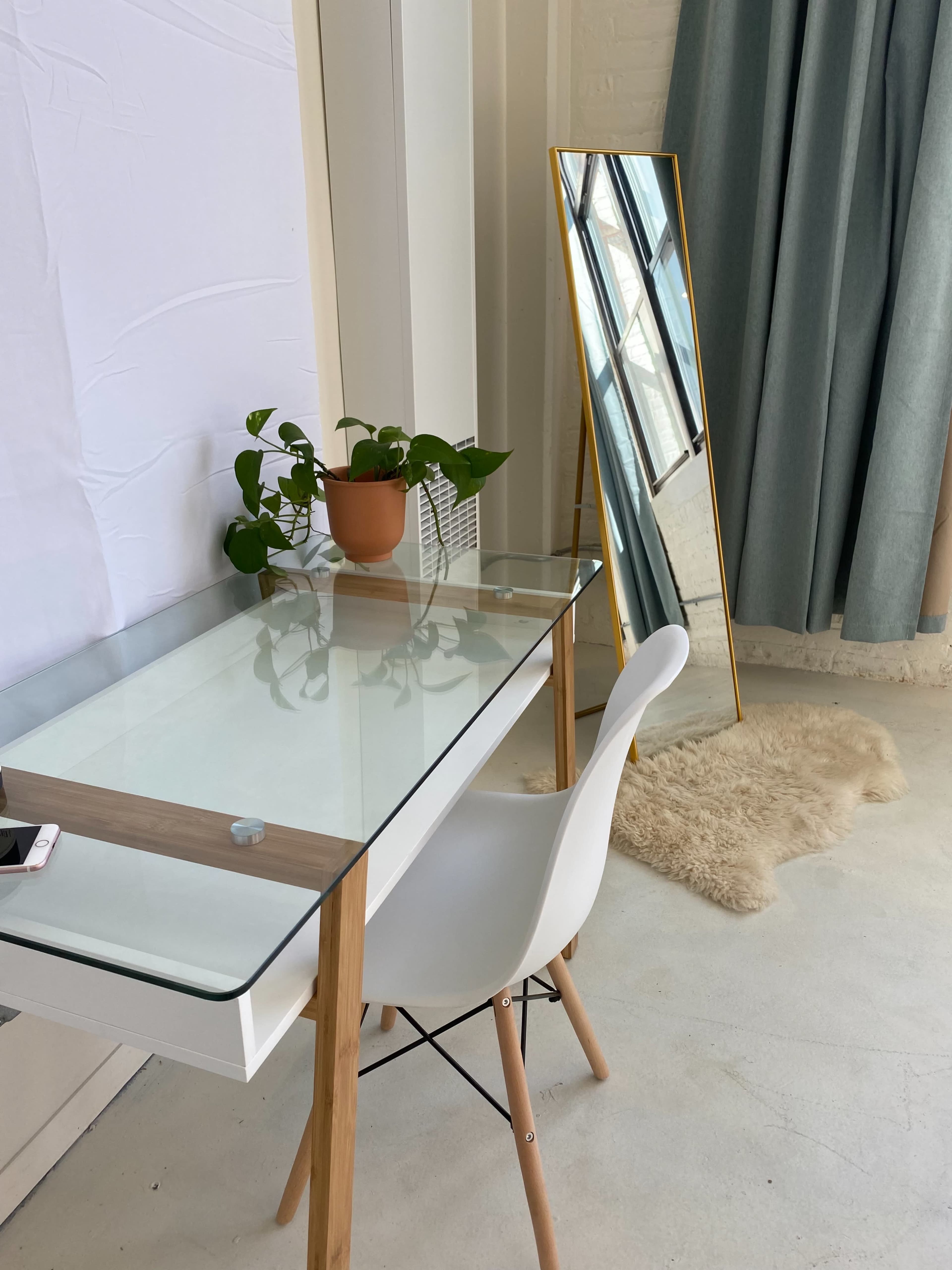 A clear-glass desk with wooden legs sits next to a potted plant and a large mirror against a wall with light curtains.