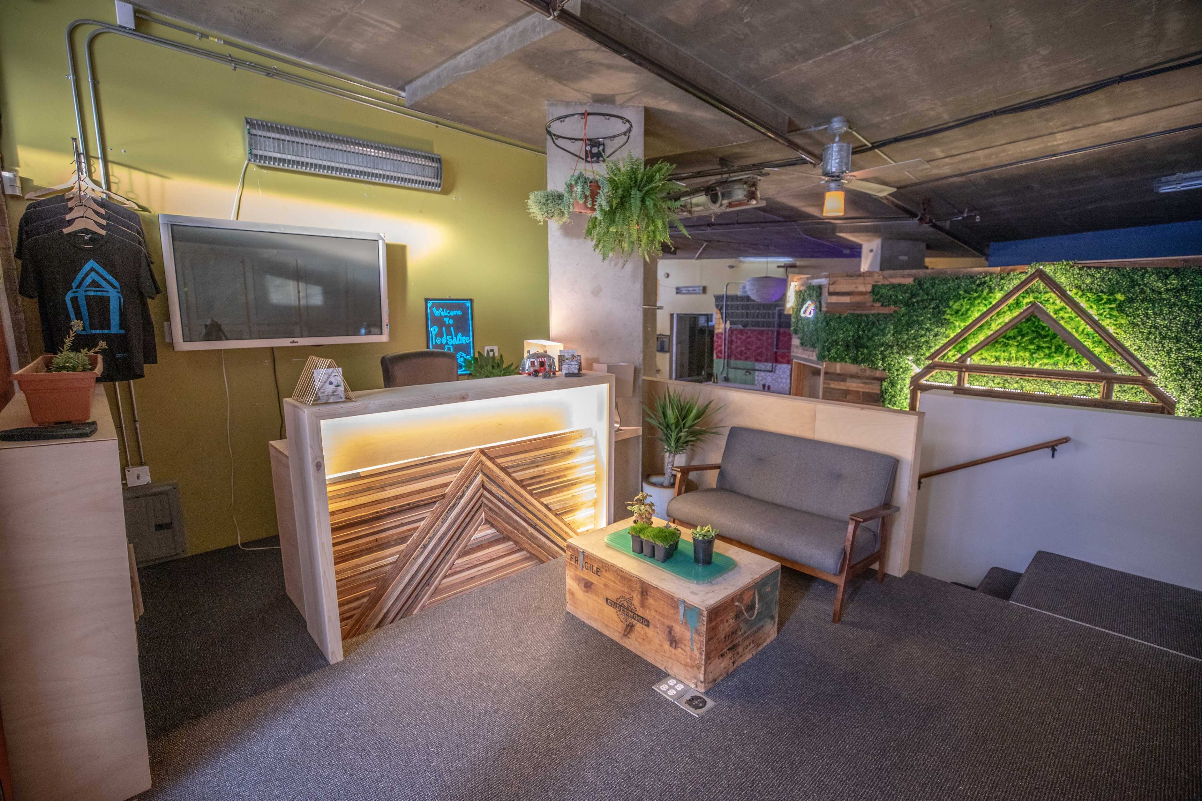 The image shows a modern reception area featuring a wooden front desk, a gray sofa, a rustic coffee table, and greenery on the walls.