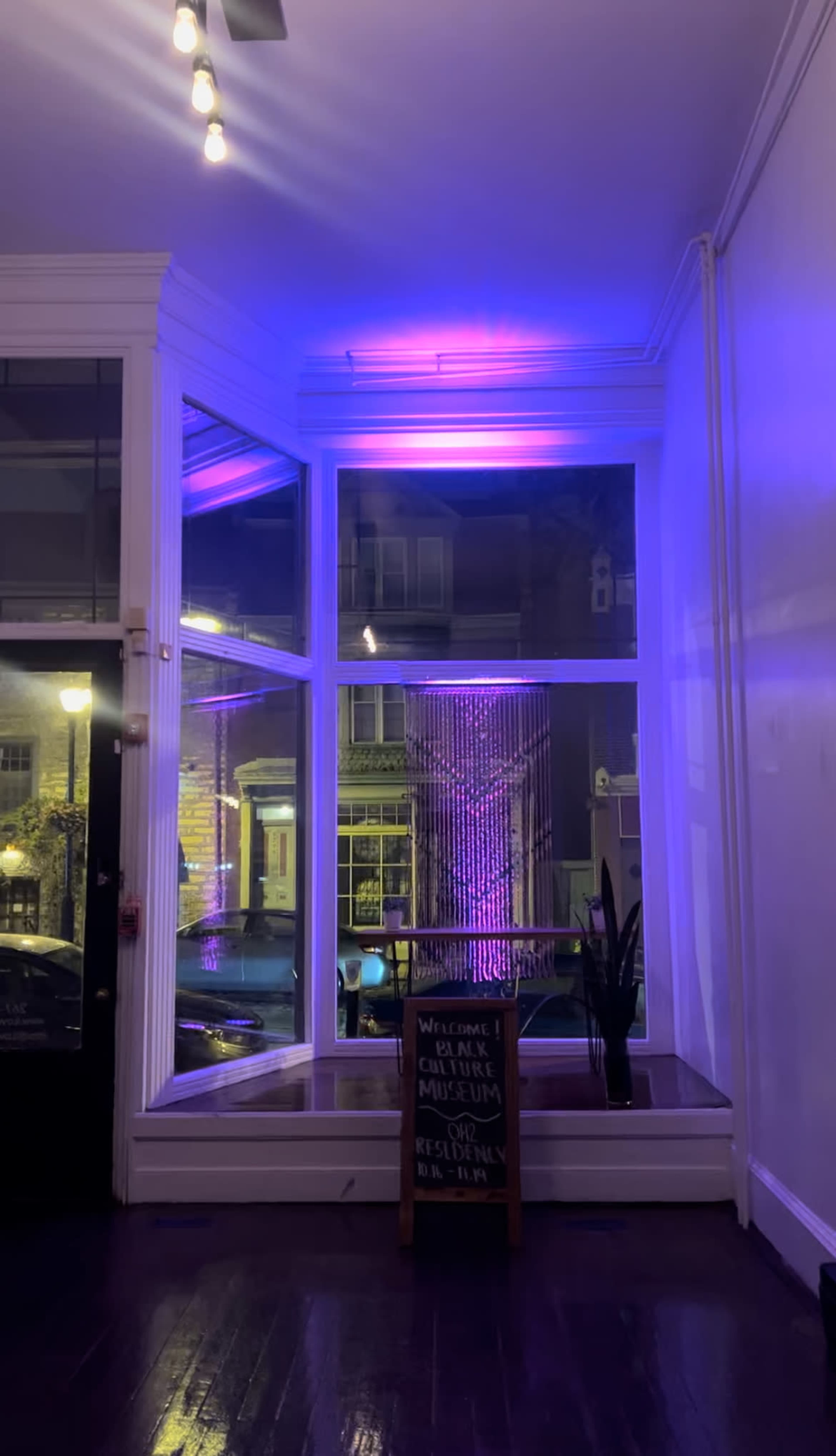The image shows an interior space with purple lighting, featuring a window that overlooks a street, and a sign on an easel welcoming visitors to a museum.