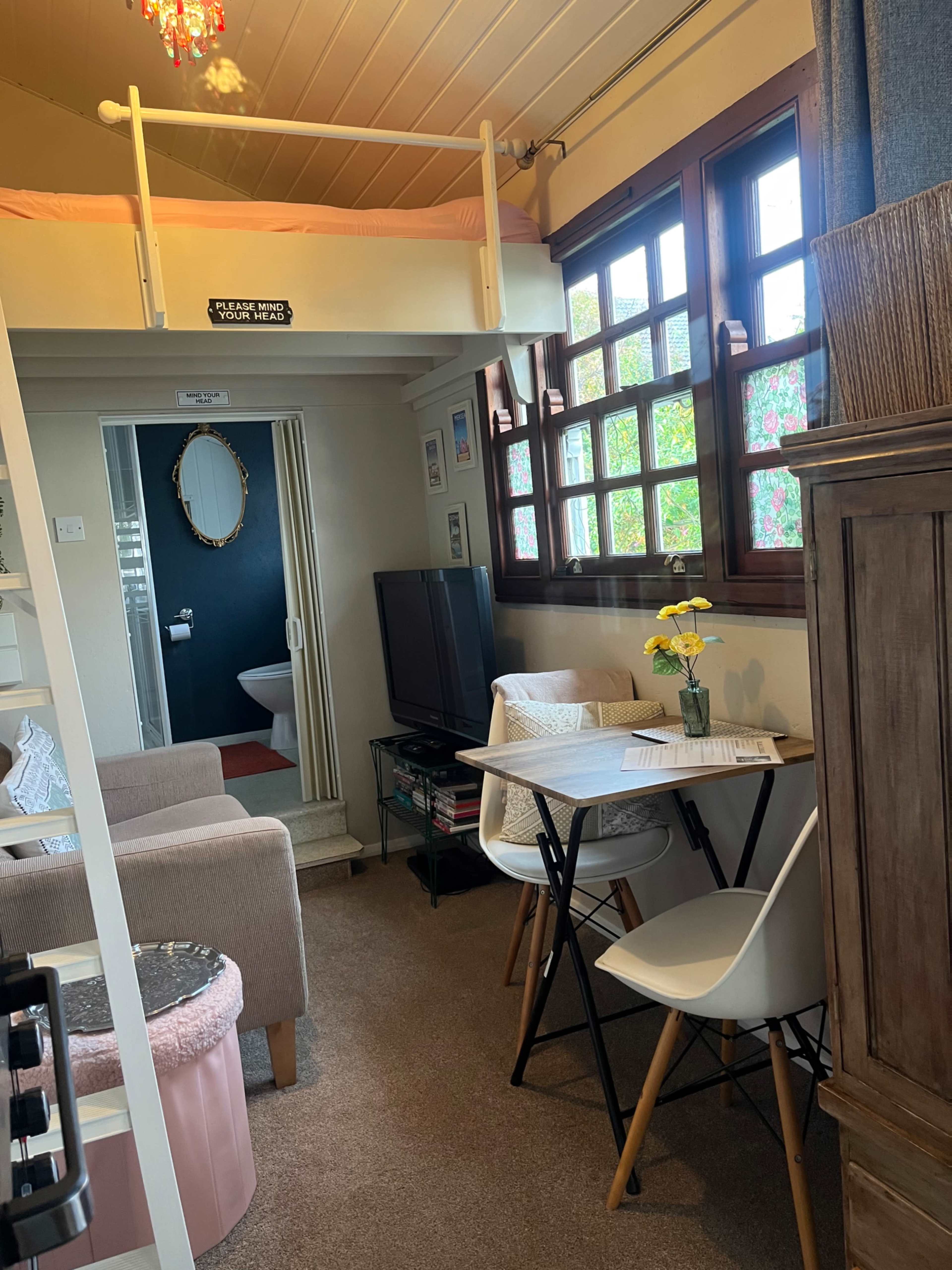 The image shows a cozy room with a lofted bed, a small seating area with a television, a table with chairs, and decorative plants near the windows.