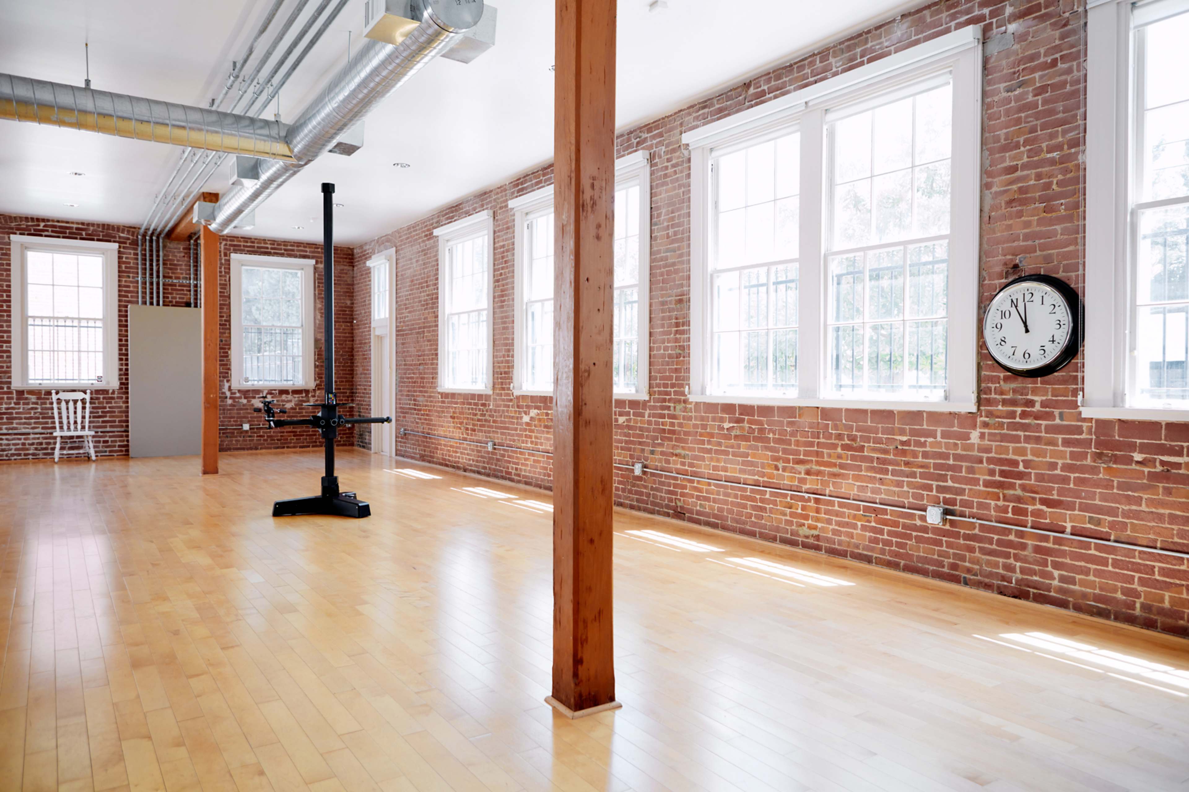 An empty room with hardwood floors, exposed brick walls, large windows, and a clock mounted on the wall.