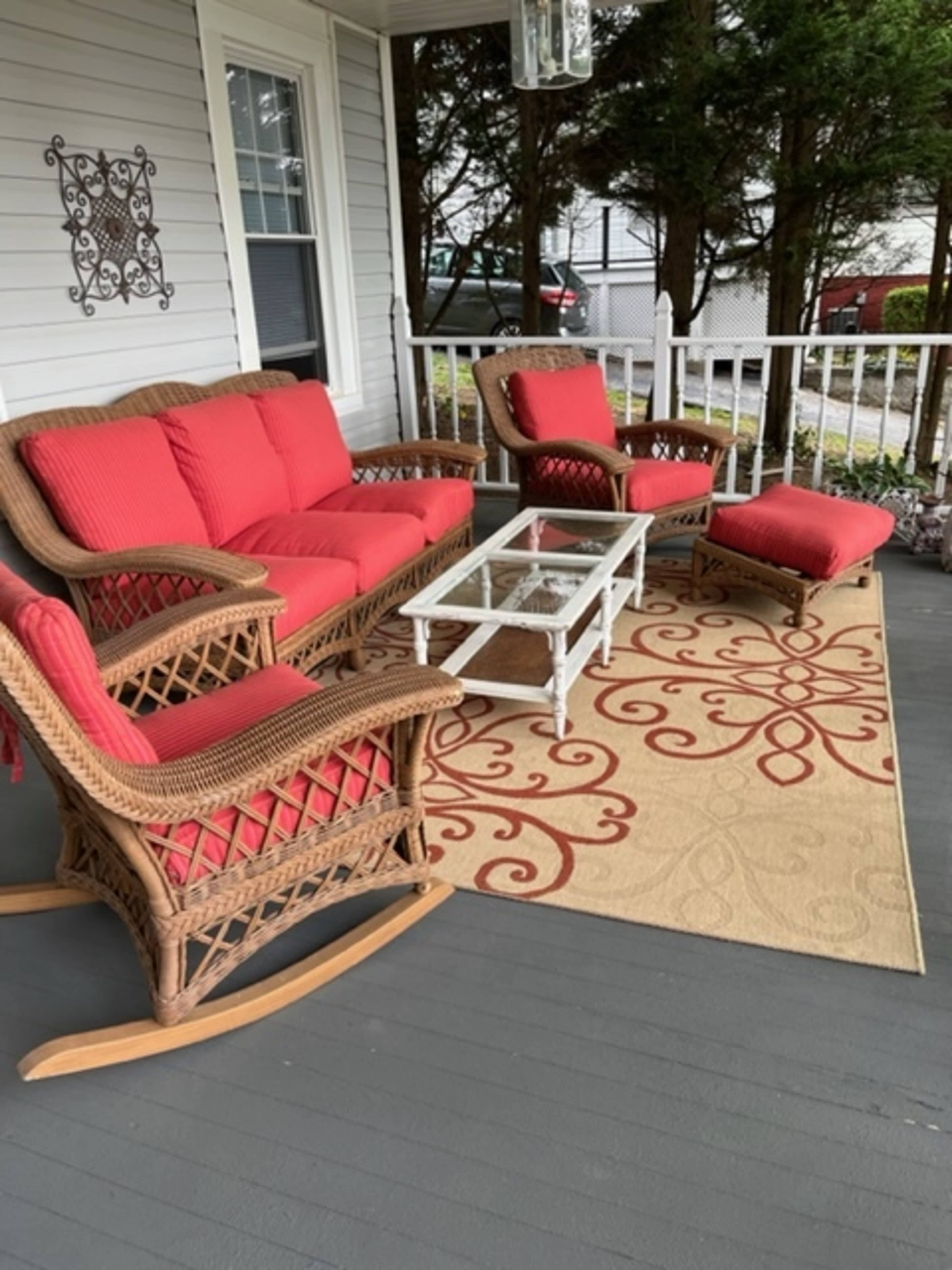 A porch features a wicker seating arrangement with red cushions, a glass coffee table, and a decorative area rug.