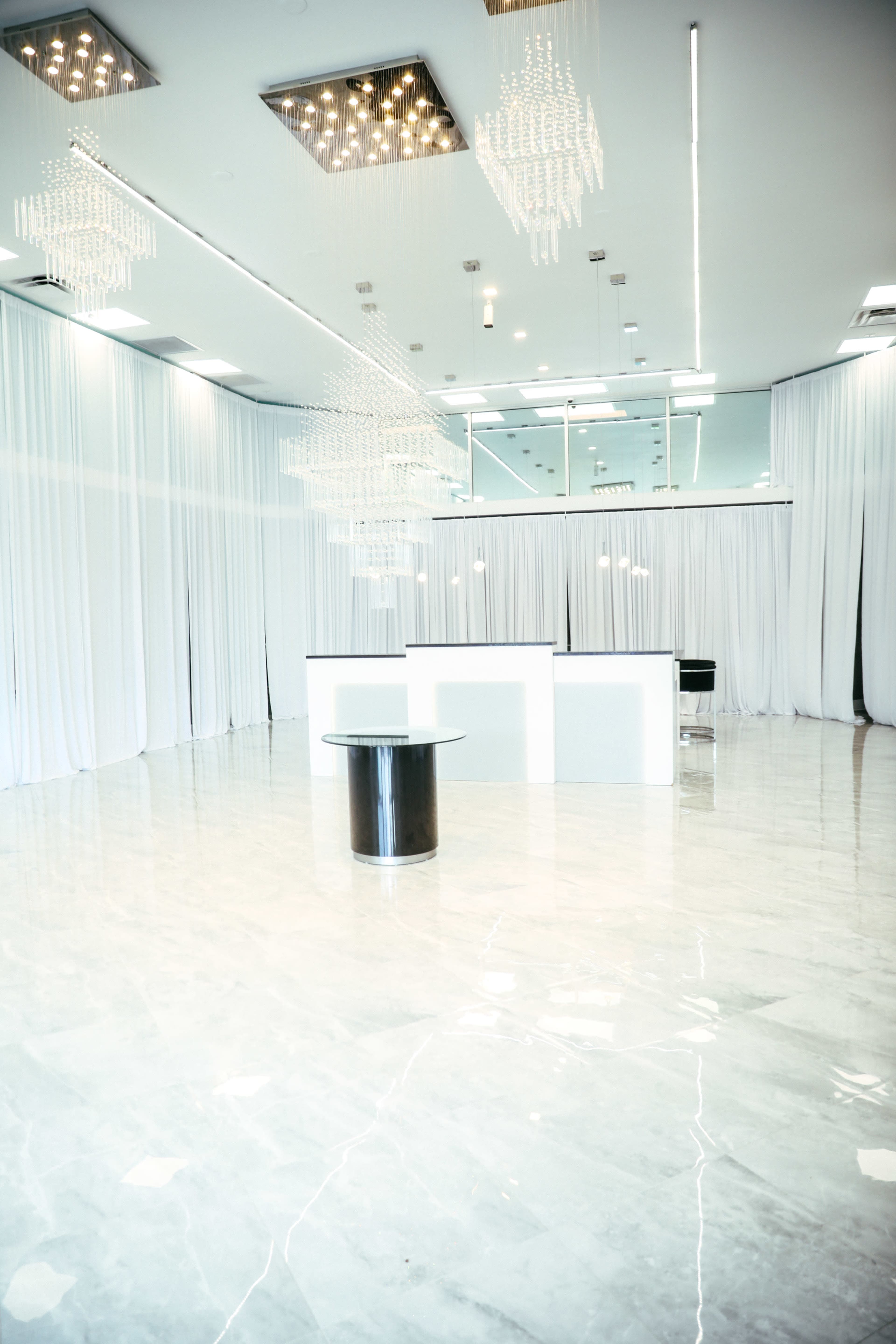 A spacious, brightly lit room features a glossy marble floor, white drapery, and modern chandeliers hanging from the ceiling.