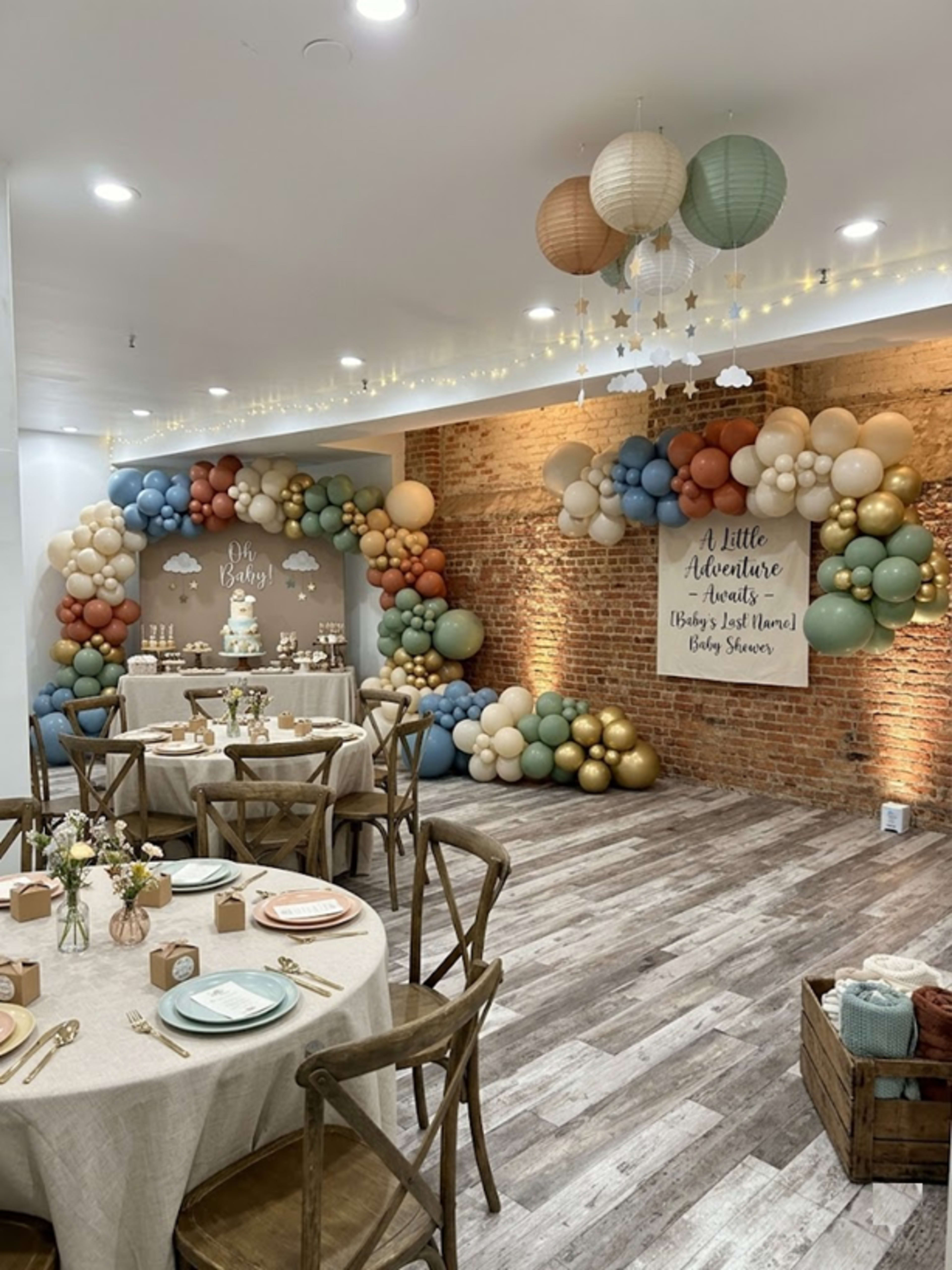 The image shows a decorated event space featuring balloon arrangements in various colors, a dessert table, and round dining tables set with floral centerpieces and tableware.