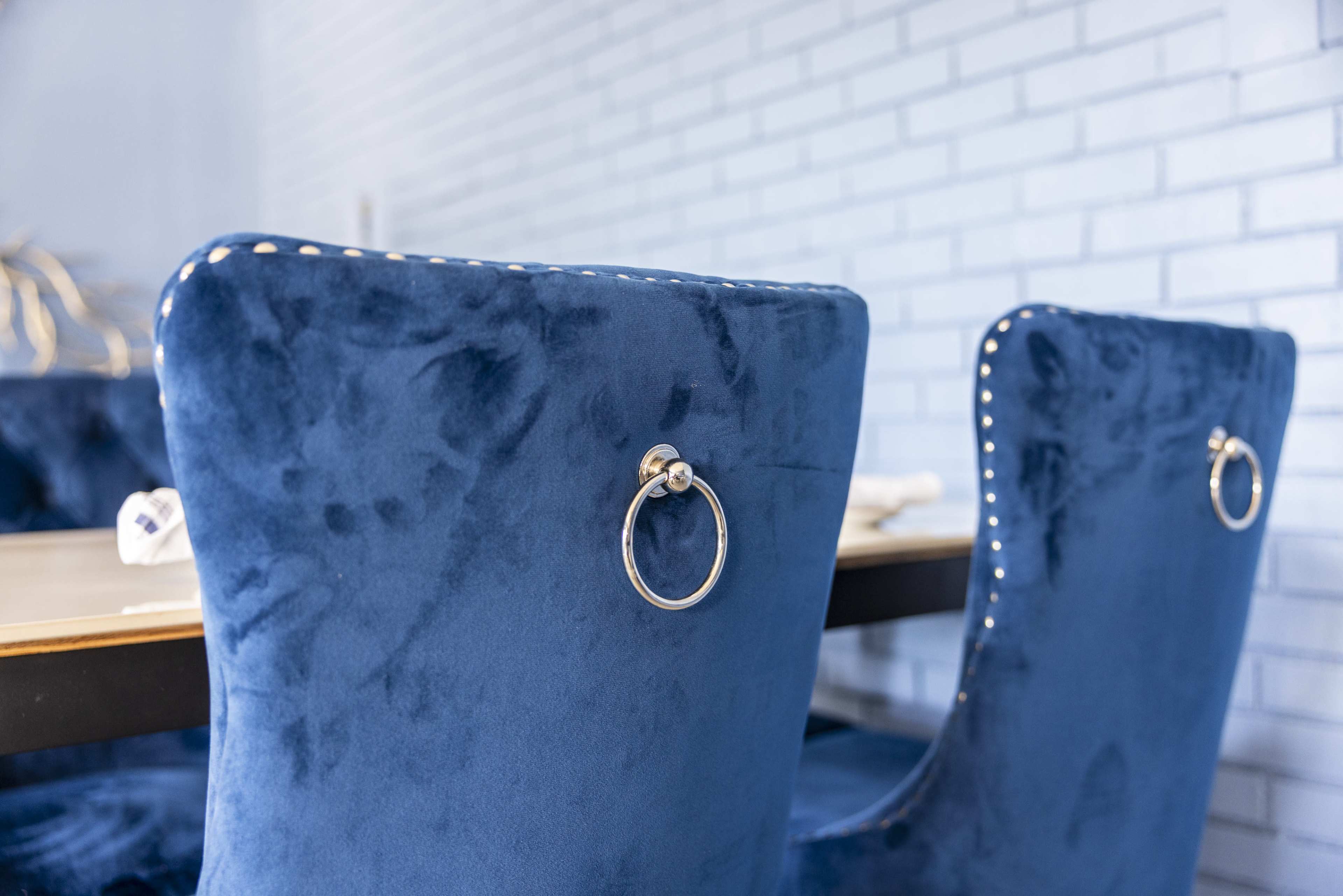 Two blue velvet dining chairs with stainless steel ring handles are positioned at a wooden table, against a light brick wall.