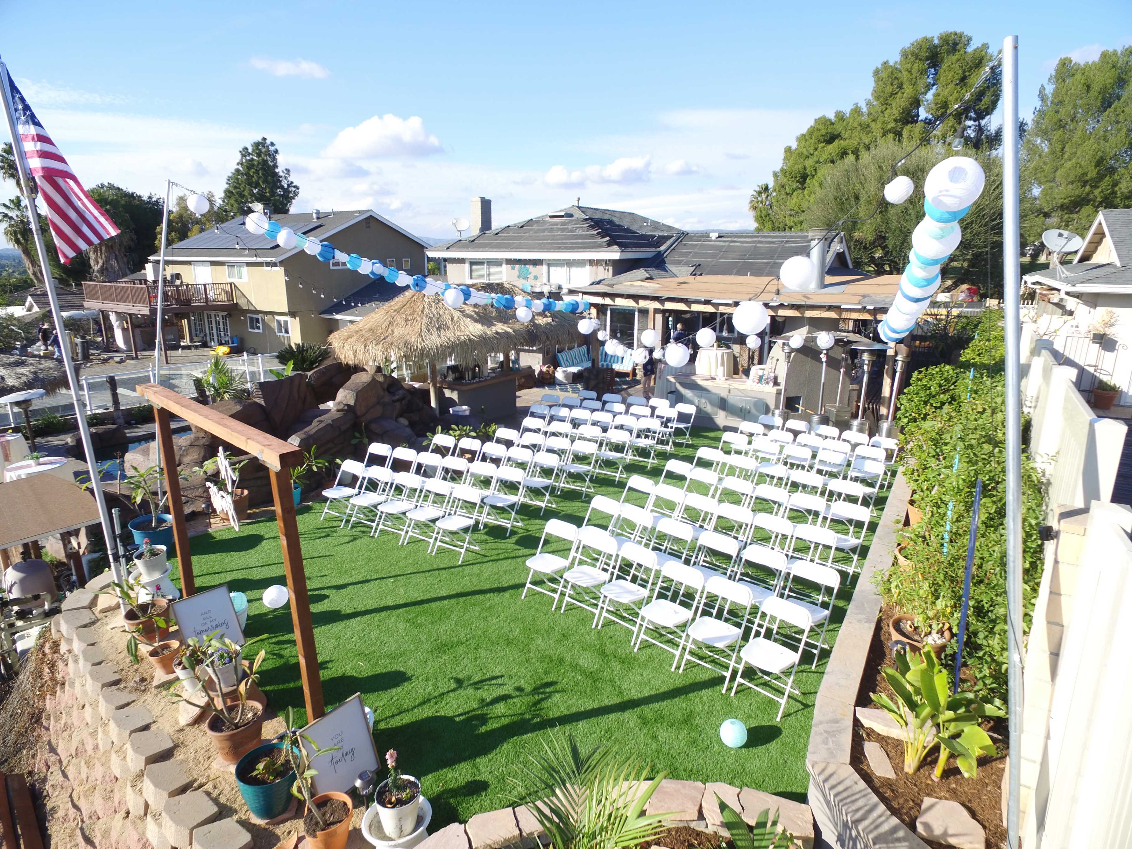 A neatly arranged outdoor seating area with white chairs set up on green grass, surrounded by decorative balloons and a thatched-roof structure.