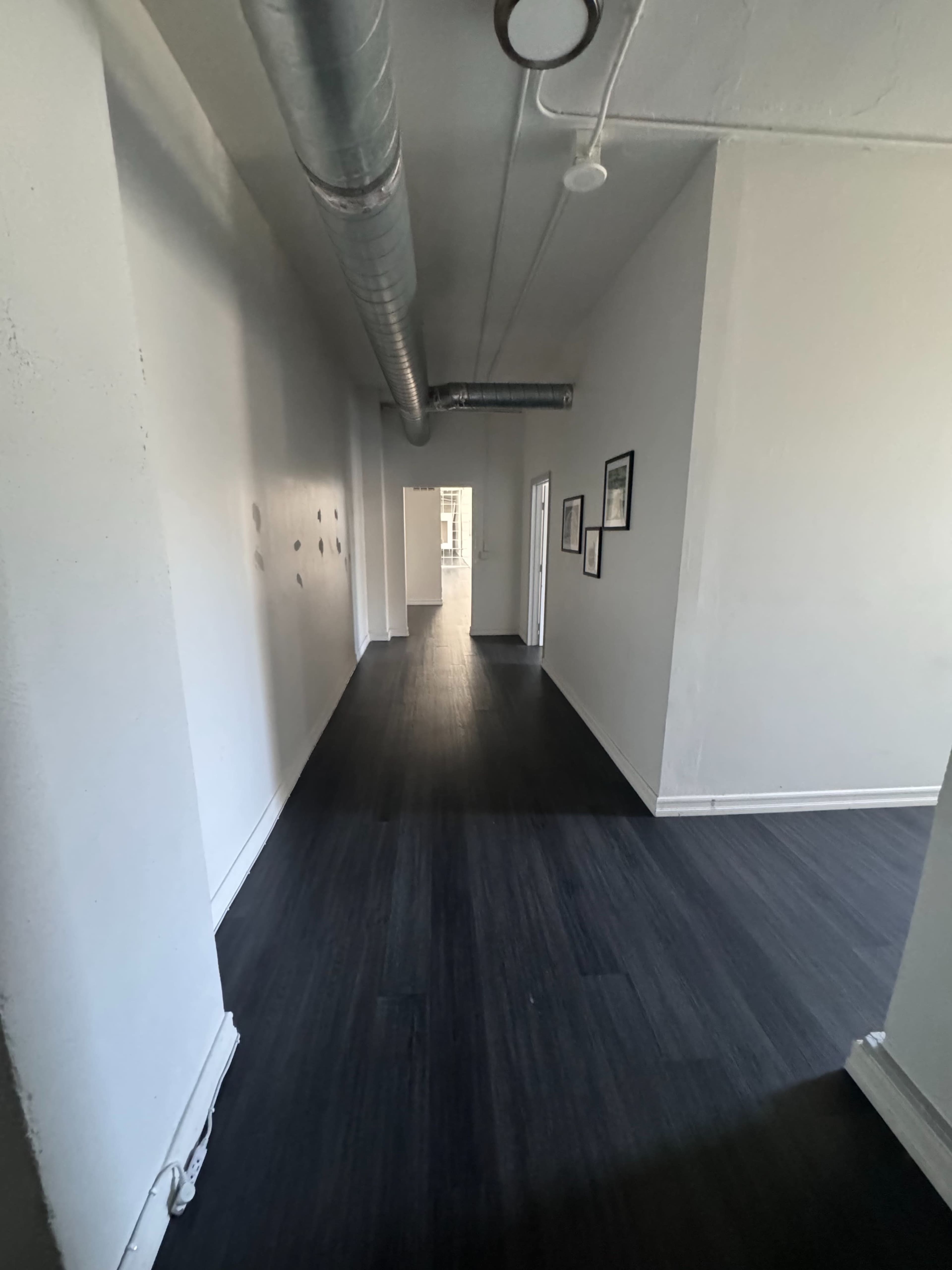 A long, narrow hallway with a dark wooden floor and white walls, featuring a metal duct running along the ceiling.