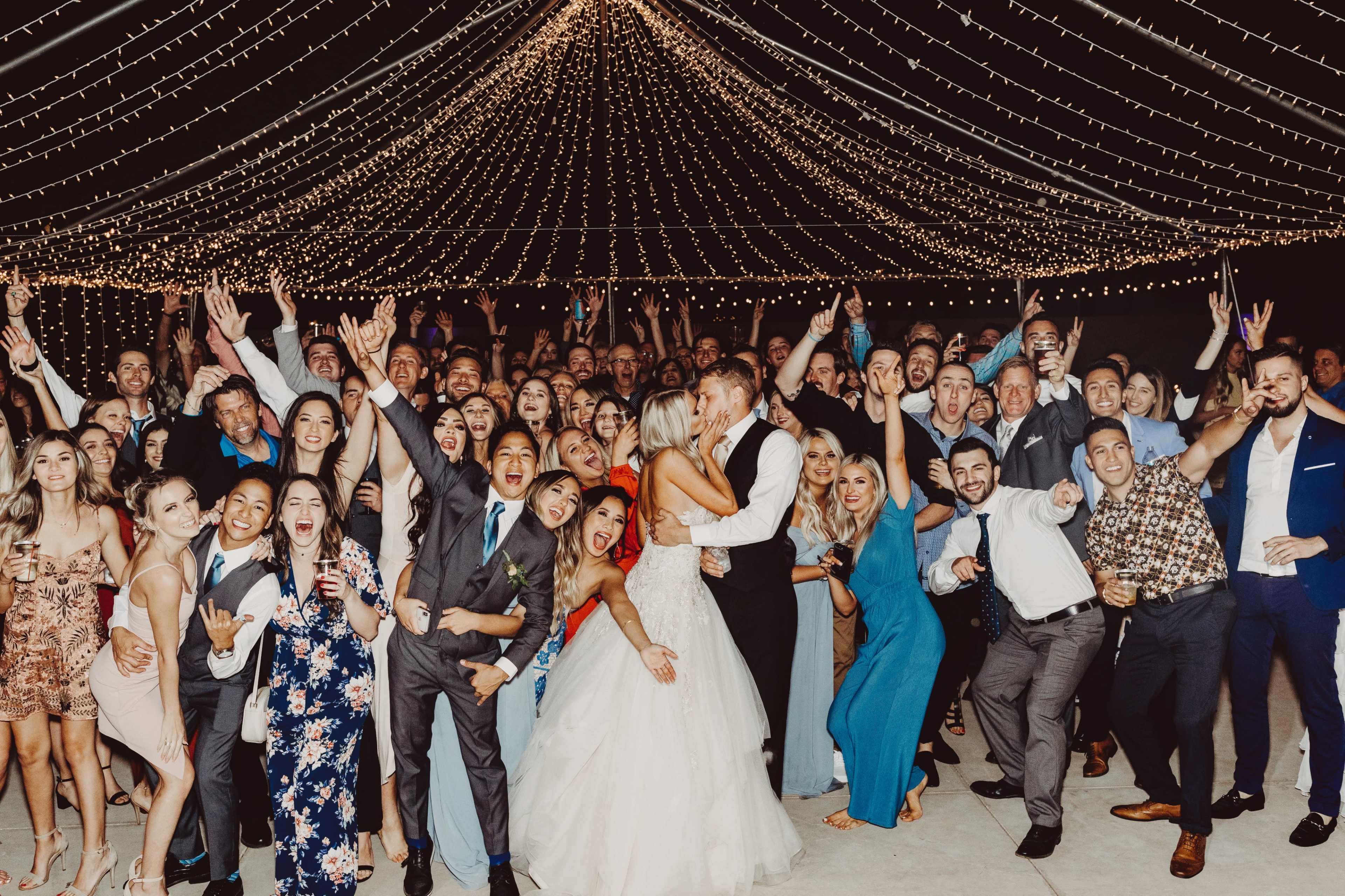 A large group of people celebrates a wedding under a canopy of fairy lights, with the bride and groom kissing at the center.