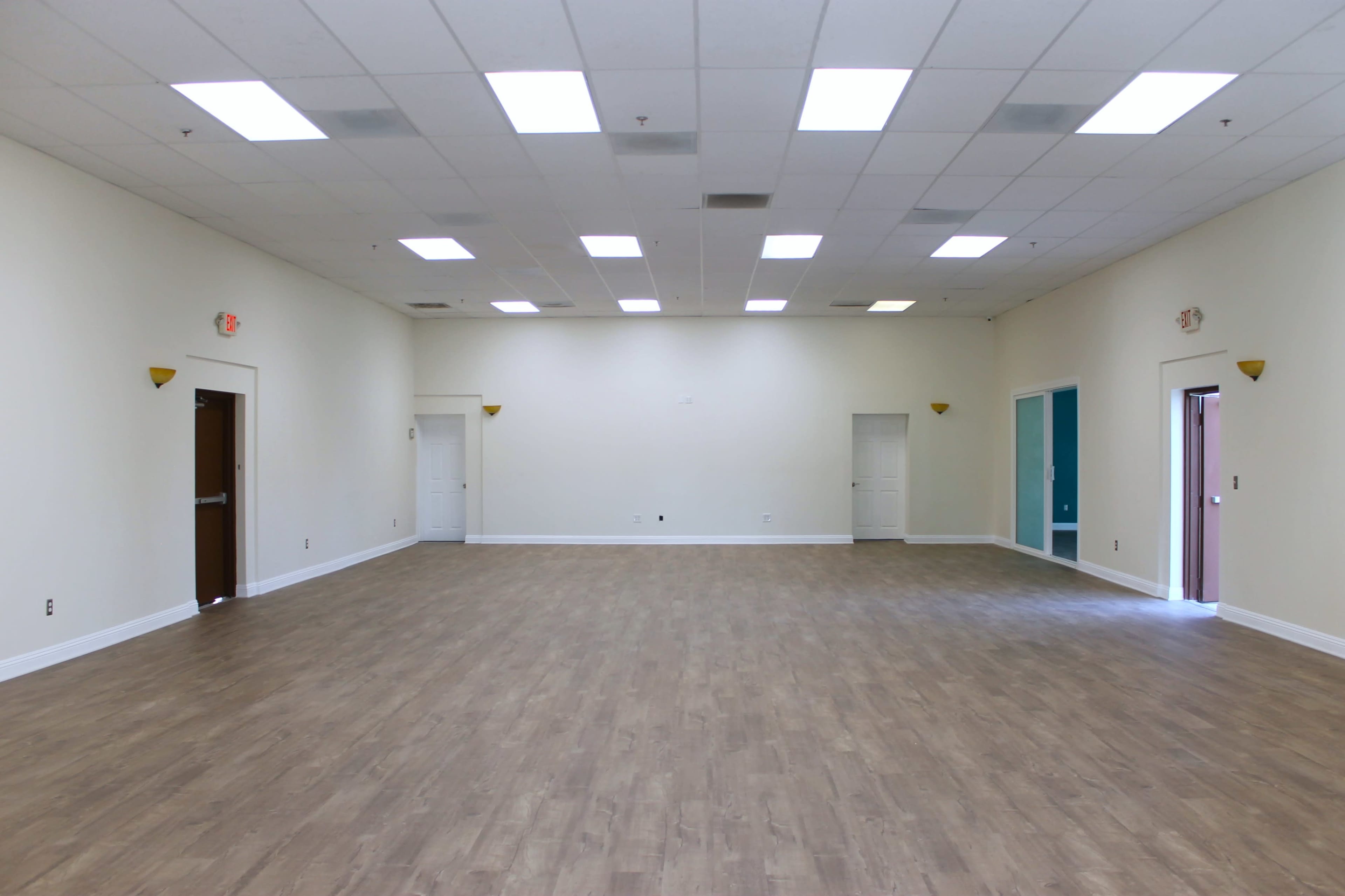 A spacious, empty room with a wooden floor, white walls, and multiple doors along the perimeter.