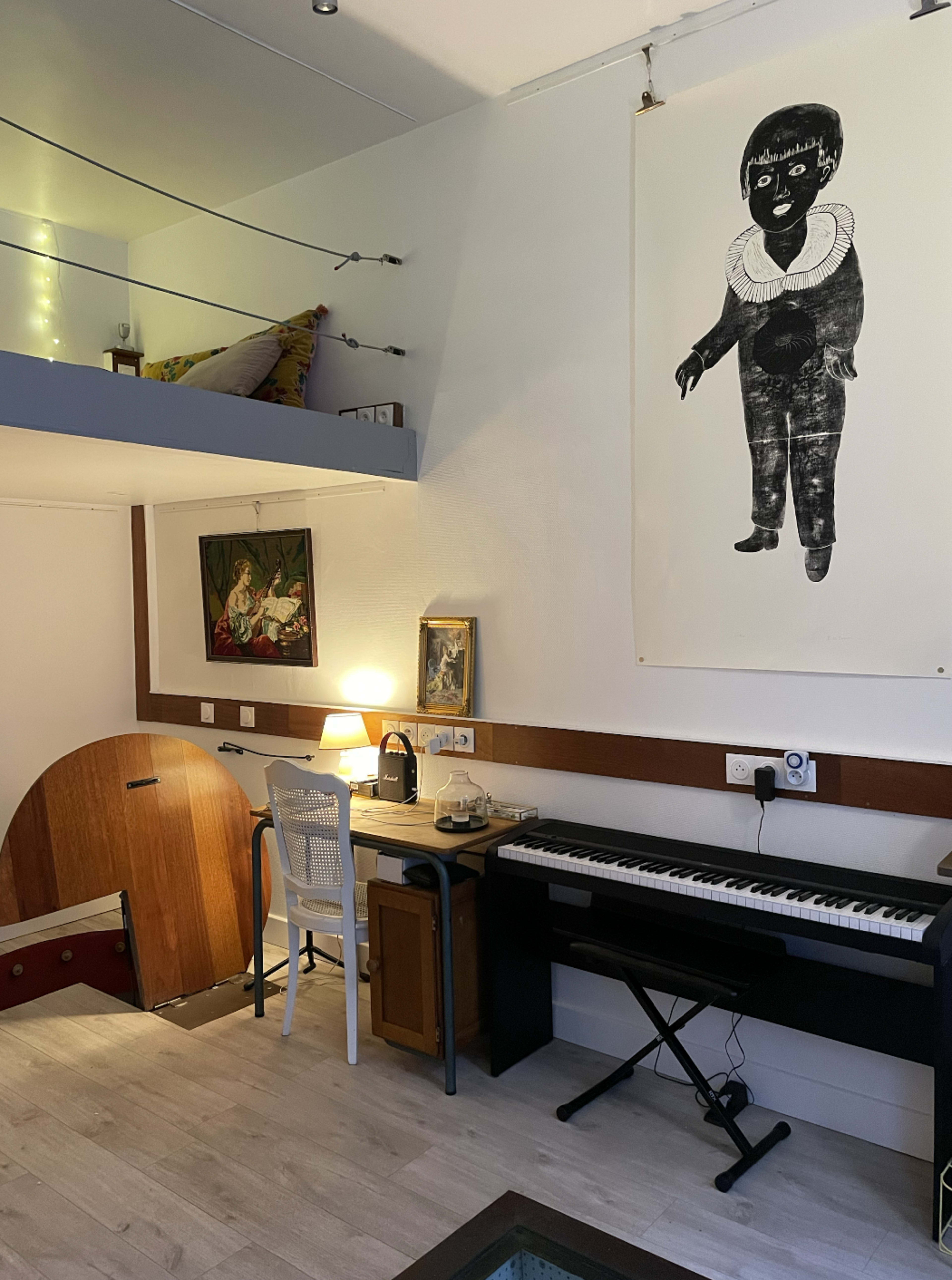 The image shows a small room with a desk and a keyboard, featuring various artworks on the walls and a staircase leading to a loft area.