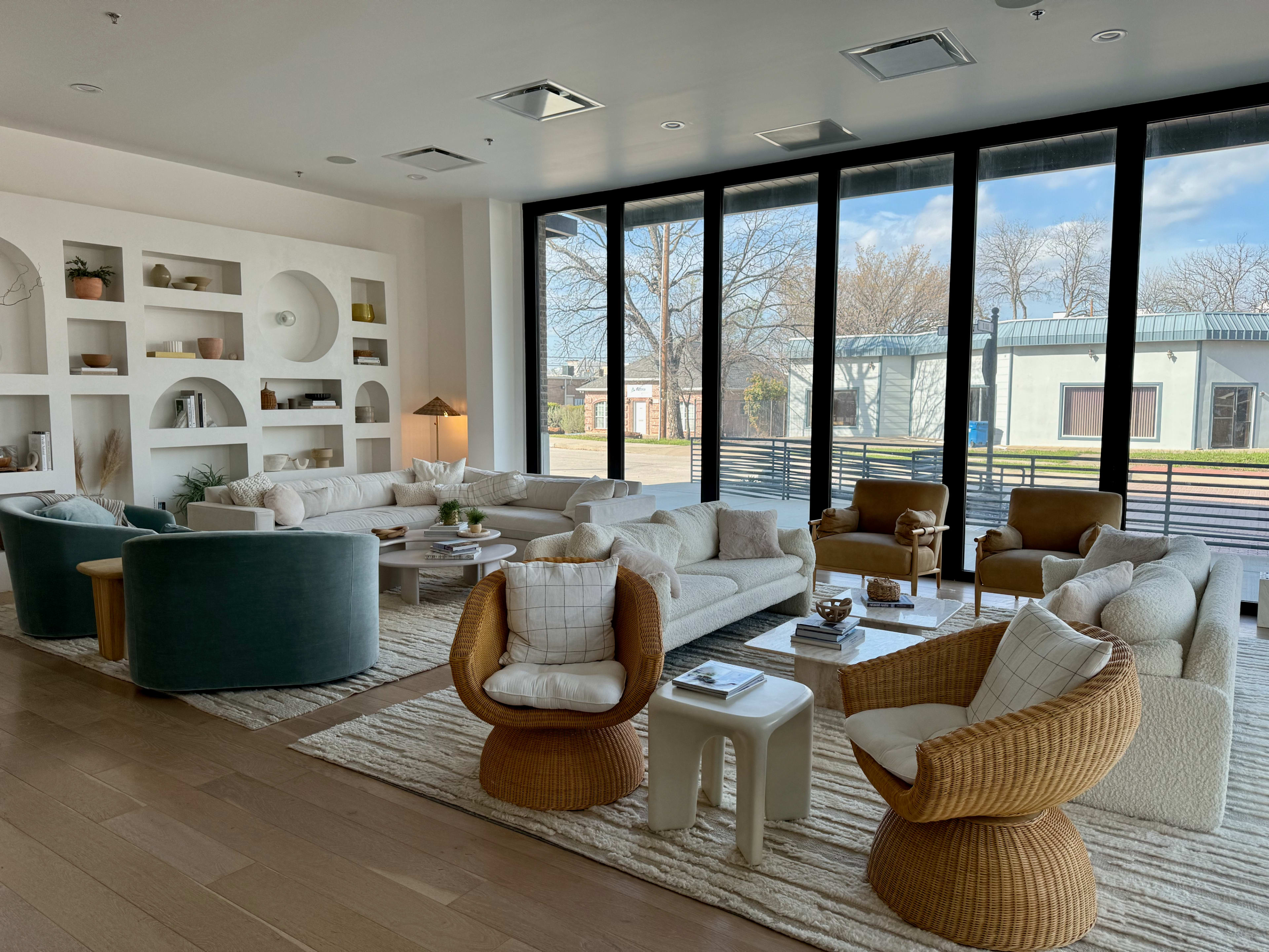A modern living room features a variety of seating options arranged around a large rug and illuminated by natural light from large windows.