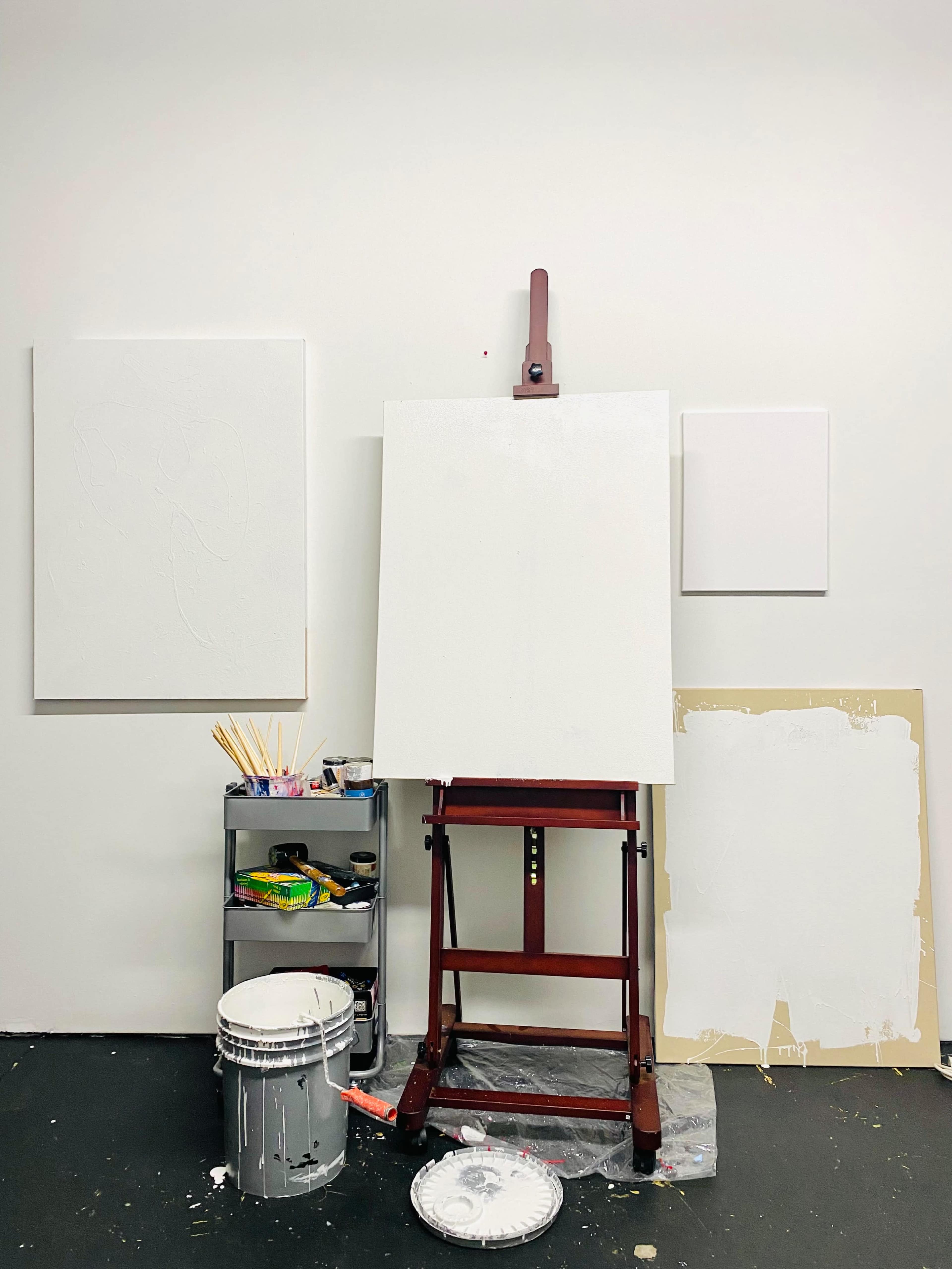 A blank canvas is set up on an easel in a room with white walls, accompanied by two empty canvases and a storage cart filled with art supplies.