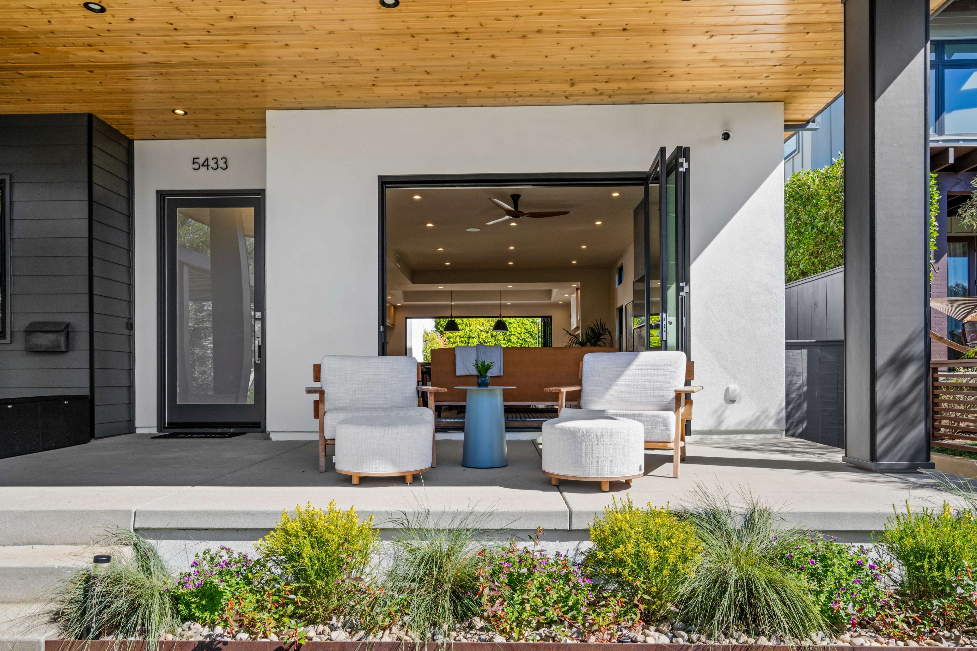 A modern porch features two chairs and a small table, surrounded by landscaped plants and an open interior visible through large glass doors.