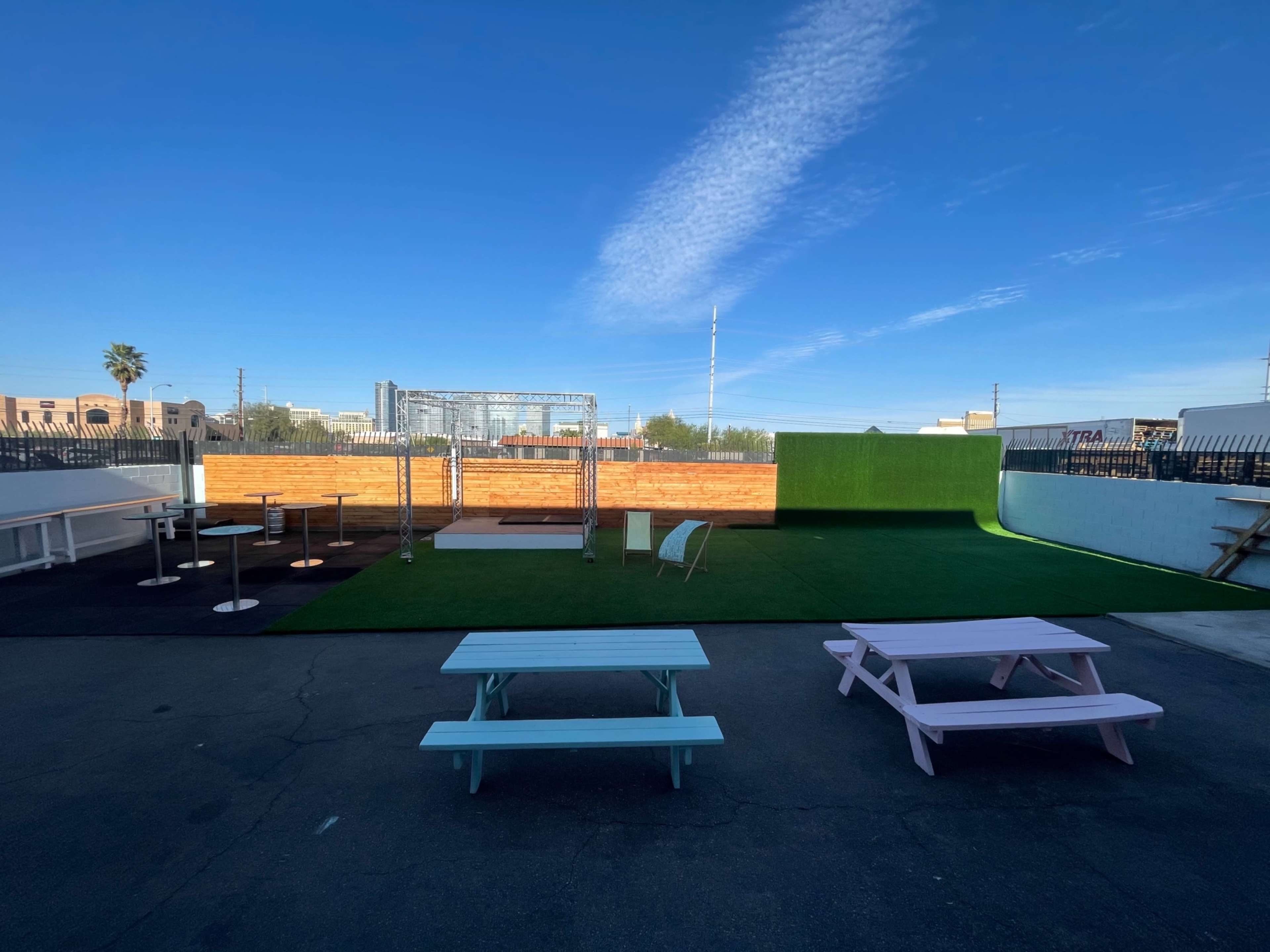 An outdoor venue with a green turf area, picnic tables, a wooden stage, and a backdrop of a blue sky.