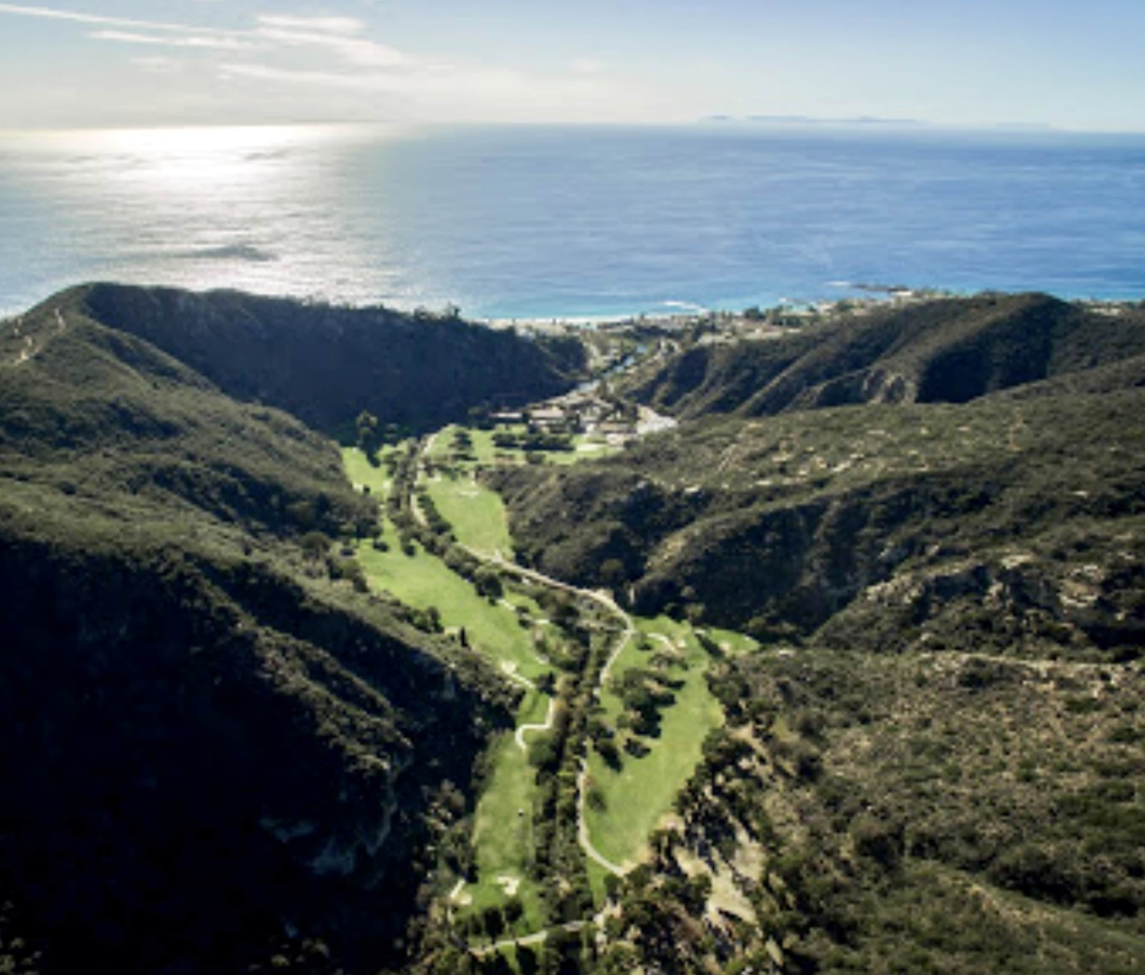 Aerial view of a golf course nestled between hills overlooking the ocean.
