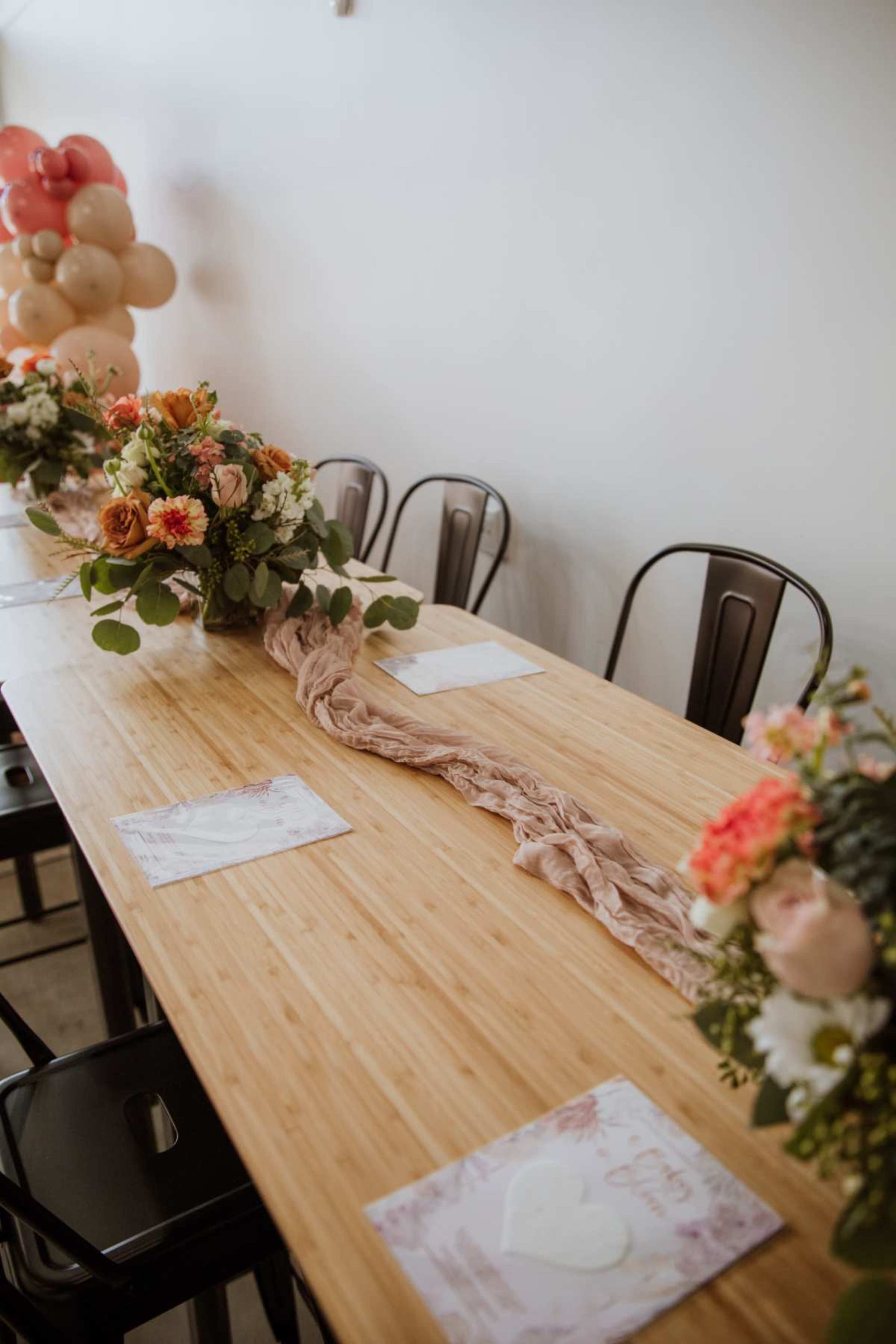 A long wooden table is set with flower arrangements on either end and decorative table runners, with empty chairs arranged around it.