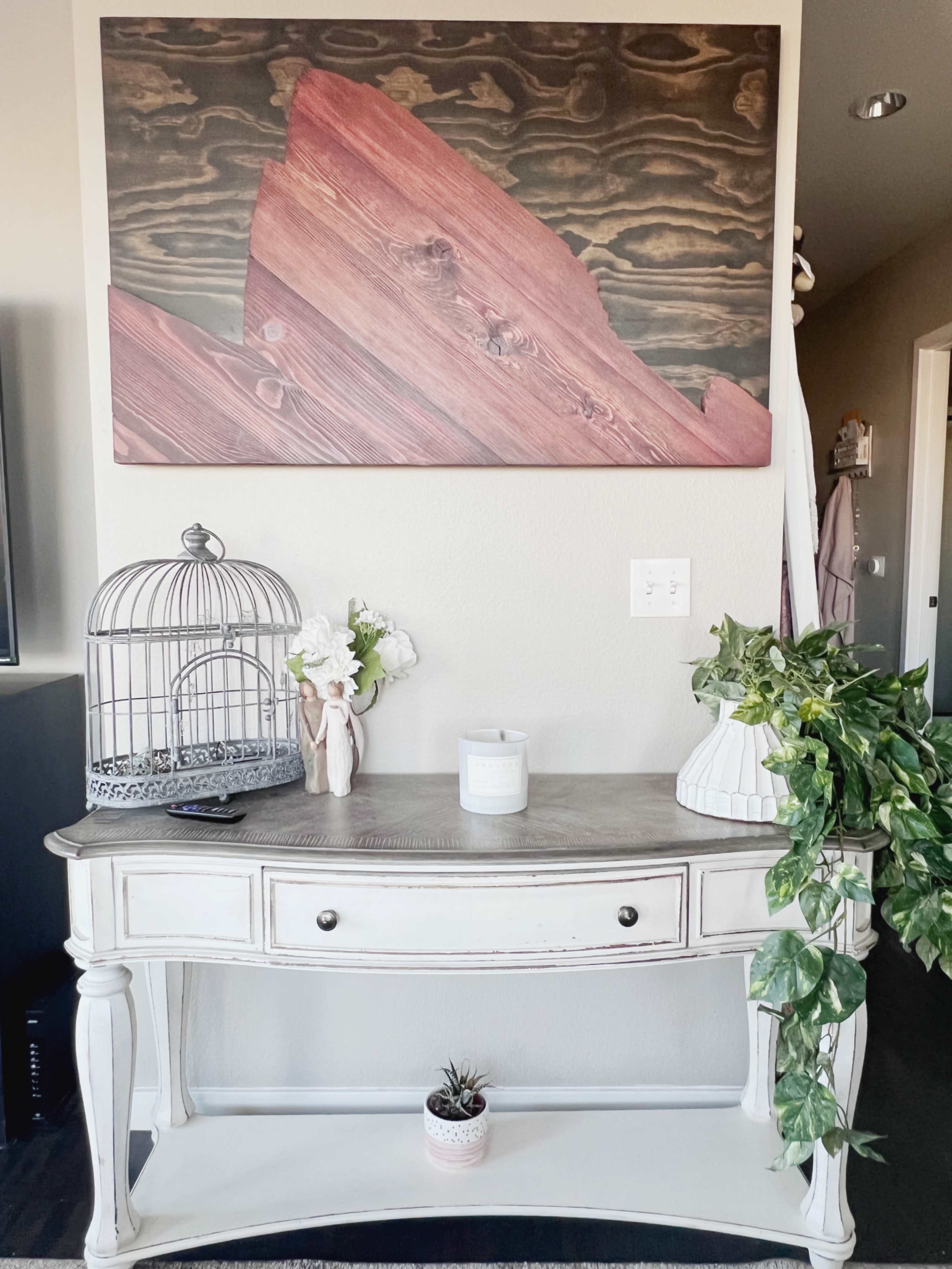 A white console table is placed against a wall adorned with a large wood-themed artwork, featuring a decorative birdcage, a vase, a candle, and a potted plant on top.