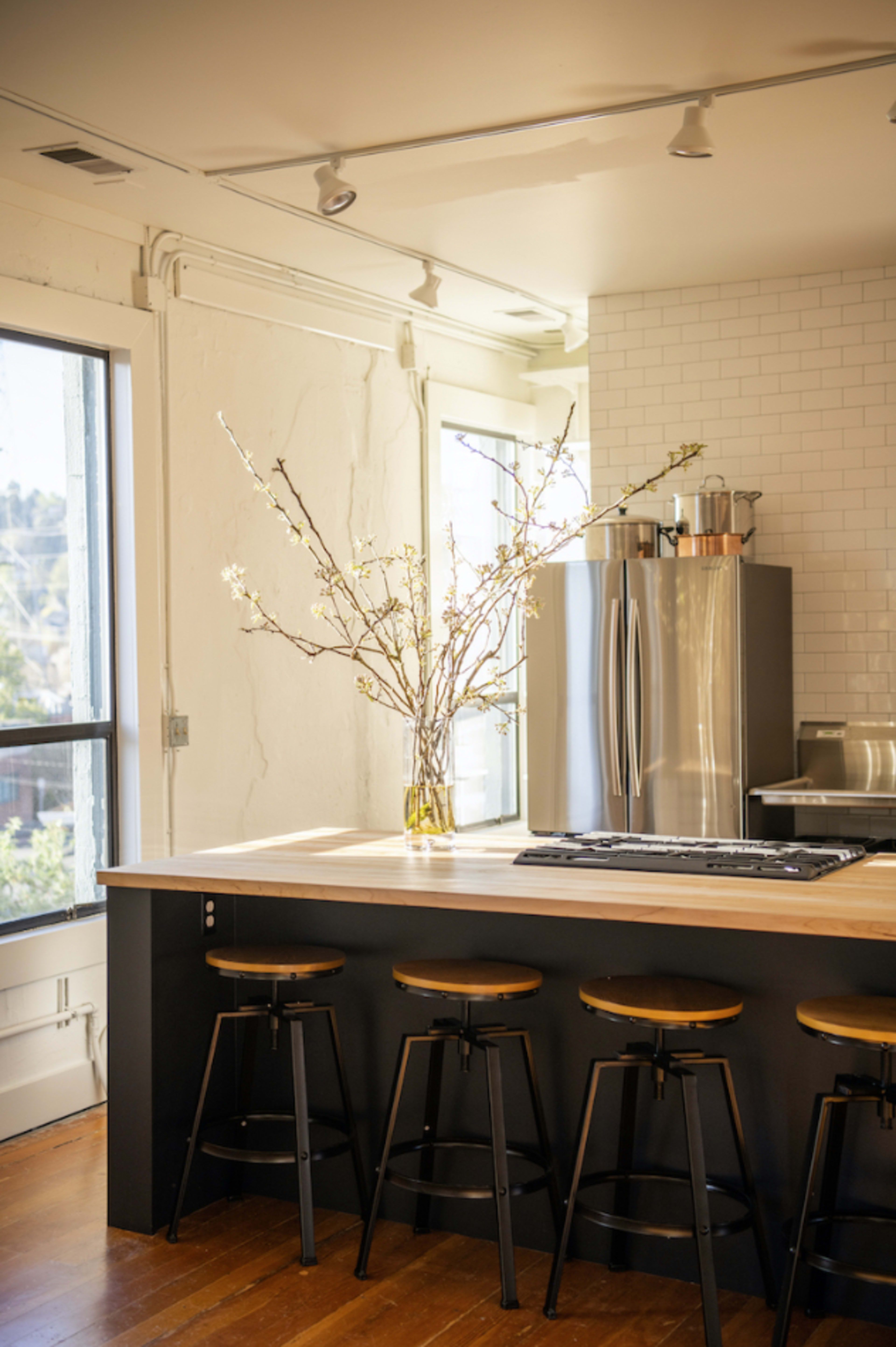 A modern kitchen features a central island with four bar stools and a vase of flowering branches on the countertop.